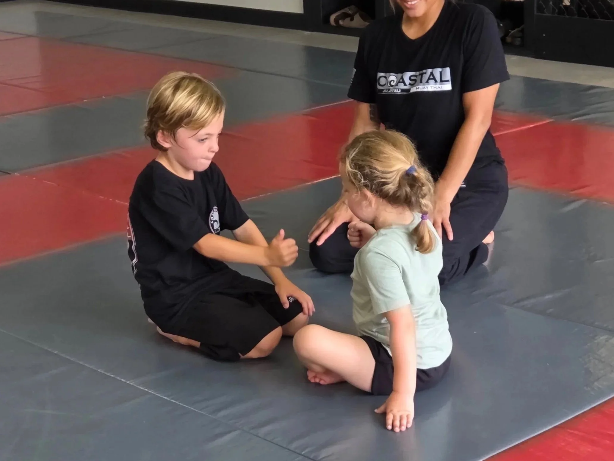 Two young children, a boy and a girl, sitting on martial arts mats during a training session, with an instructor kneeling behind them on the mats.