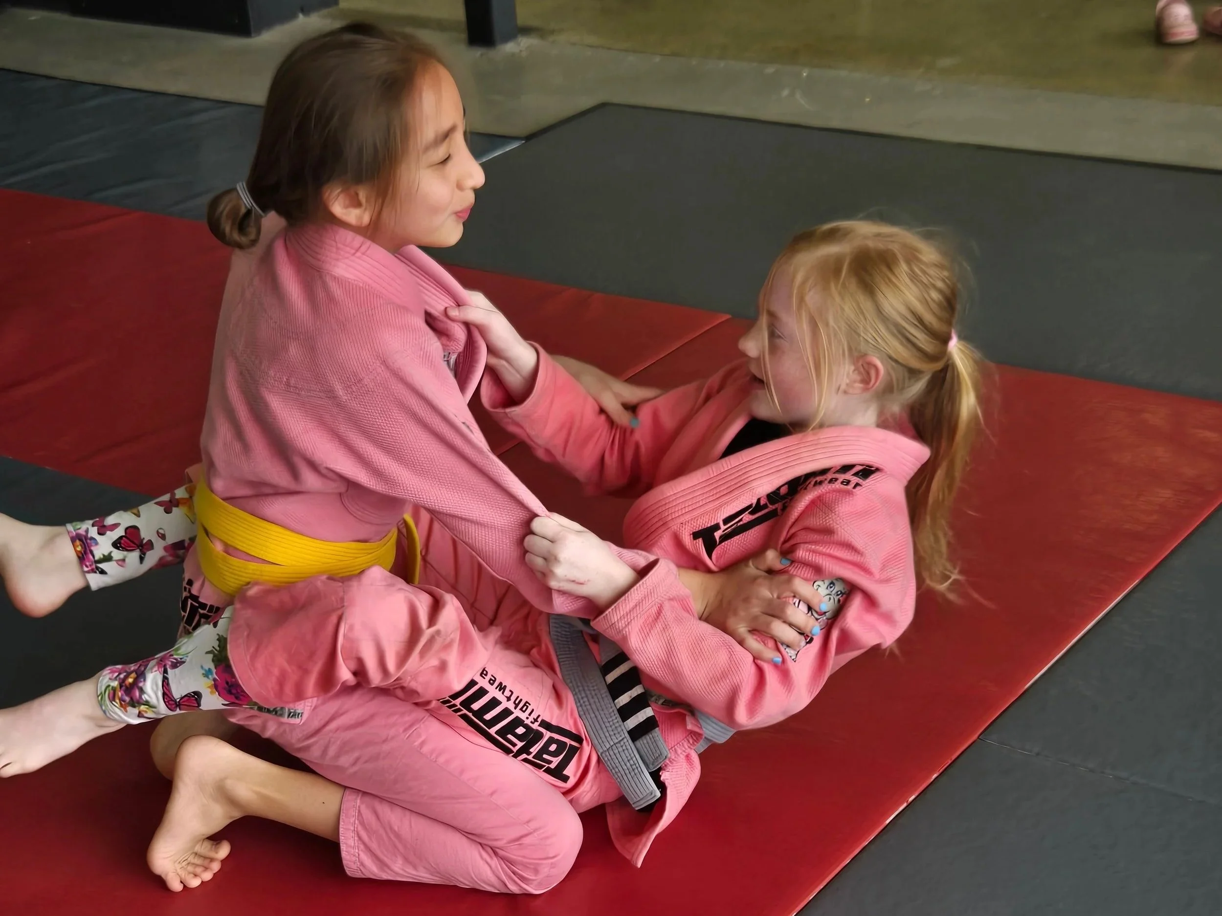 Two girls practicing Brazilian Jiu-Jitsu on a red and black mat, wearing pink gis and yellow belts, engaging in a playful sparring session.