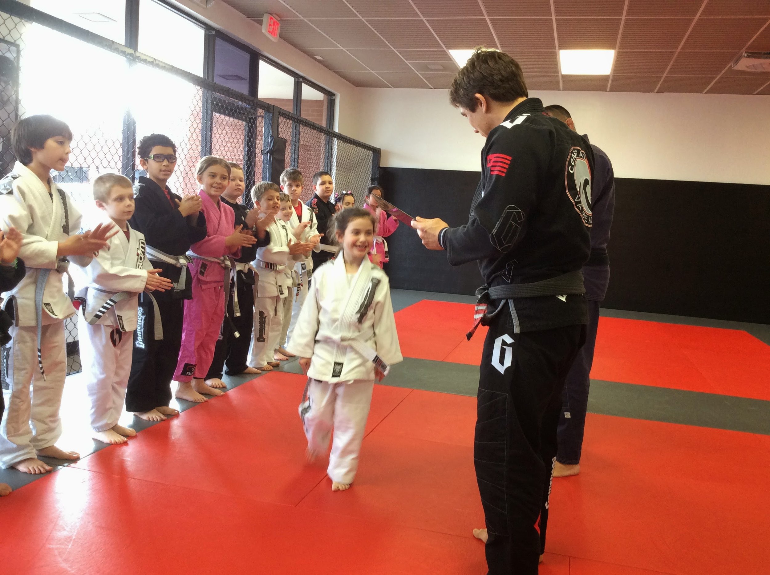 Young girl receiving an award or certificate from a martial arts instructor in a martial arts gym, with children in martial arts uniforms clapping and watching.