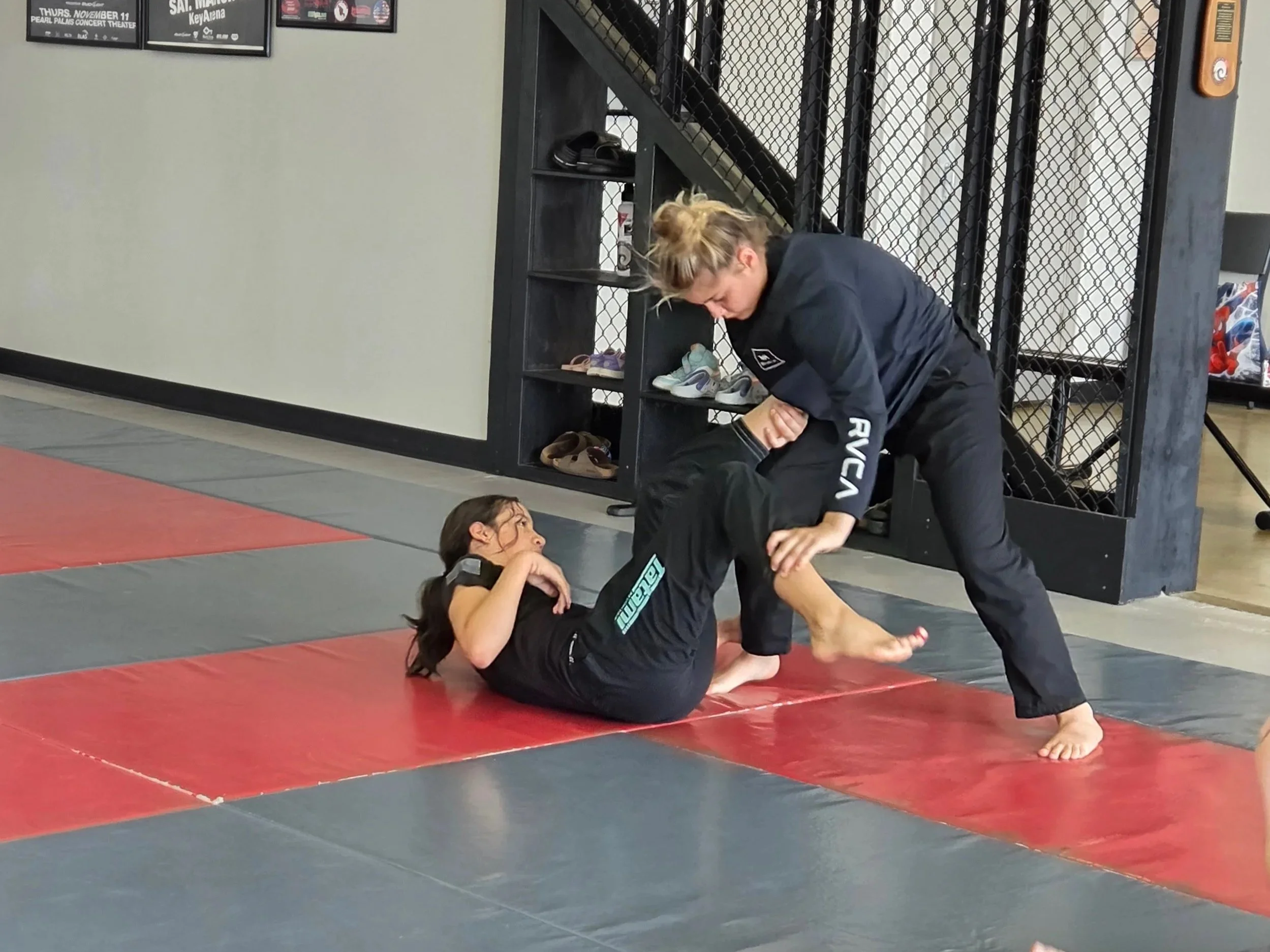 Two women practicing Brazilian Jiu-Jitsu on a mat, with one lying on her back and the other standing, gripping her opponent's arm in a grappling move.