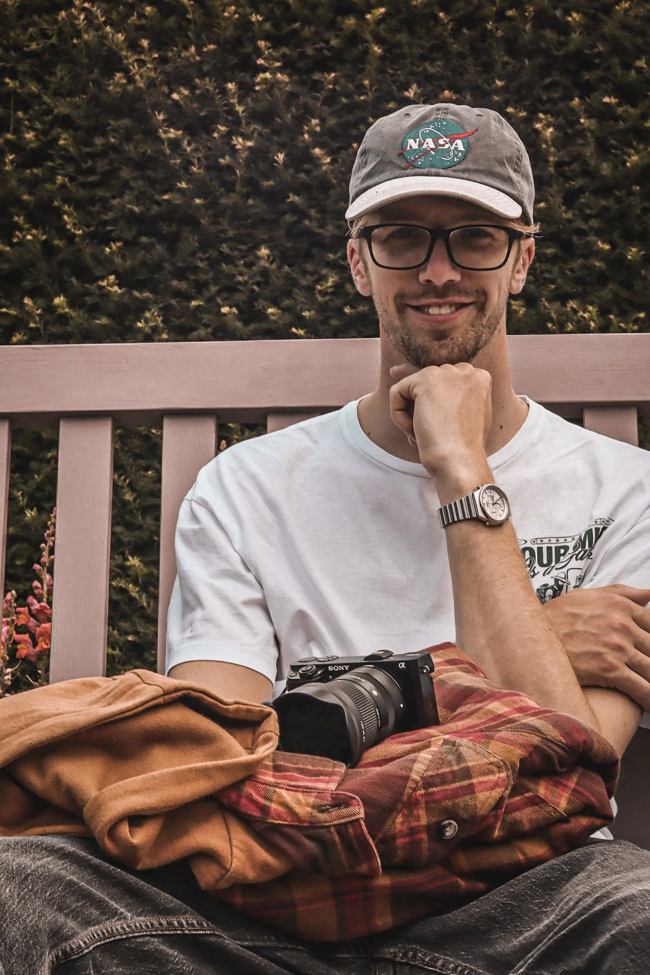A man with glasses, a gray NASA cap, a white T-shirt, and a watch, sitting outdoors on a pink bench. A camera and a brown plaid shirt are placed on his lap, with a background of dense greenery.