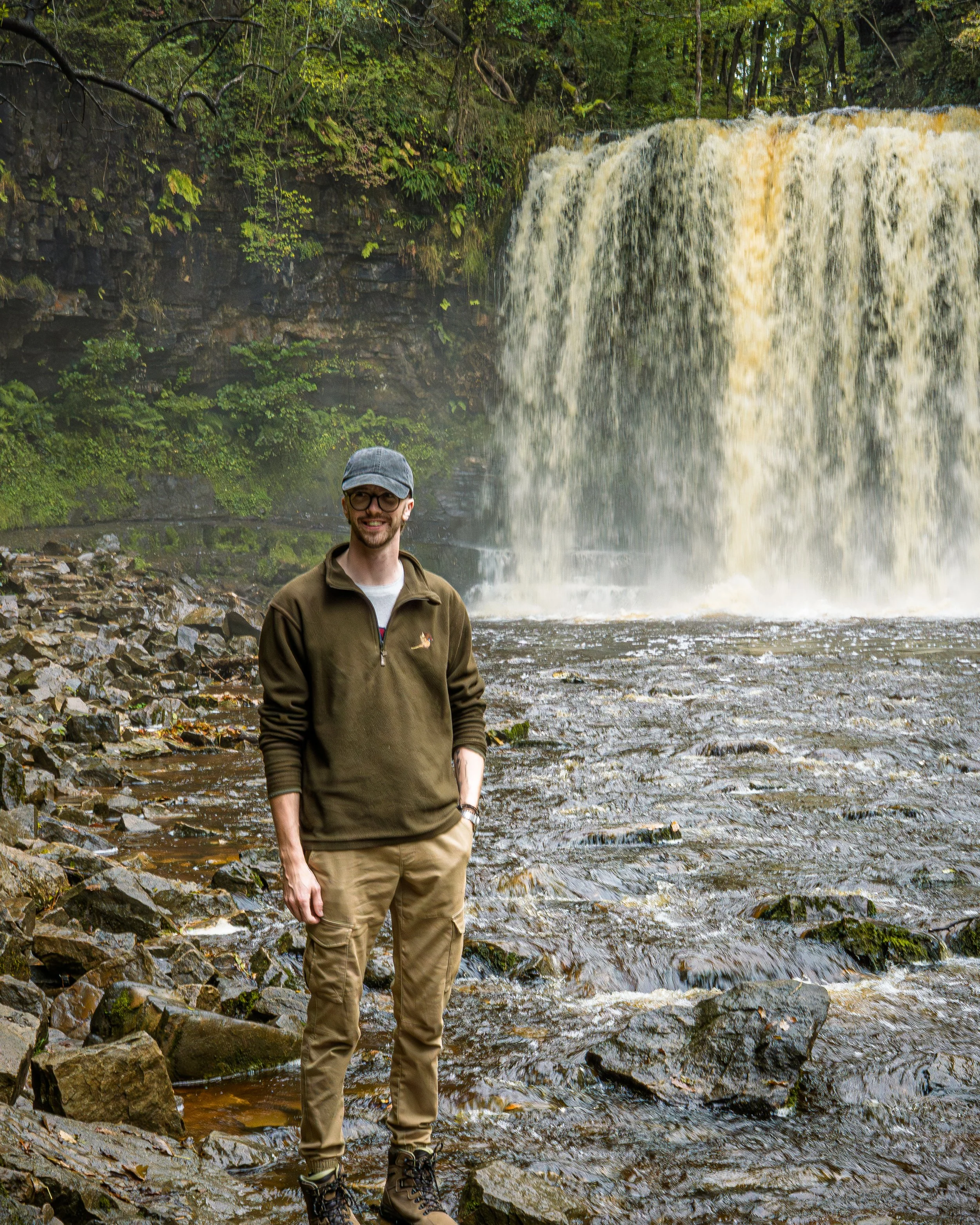 A man standing on rocks near a waterfall in a lush, green forest.