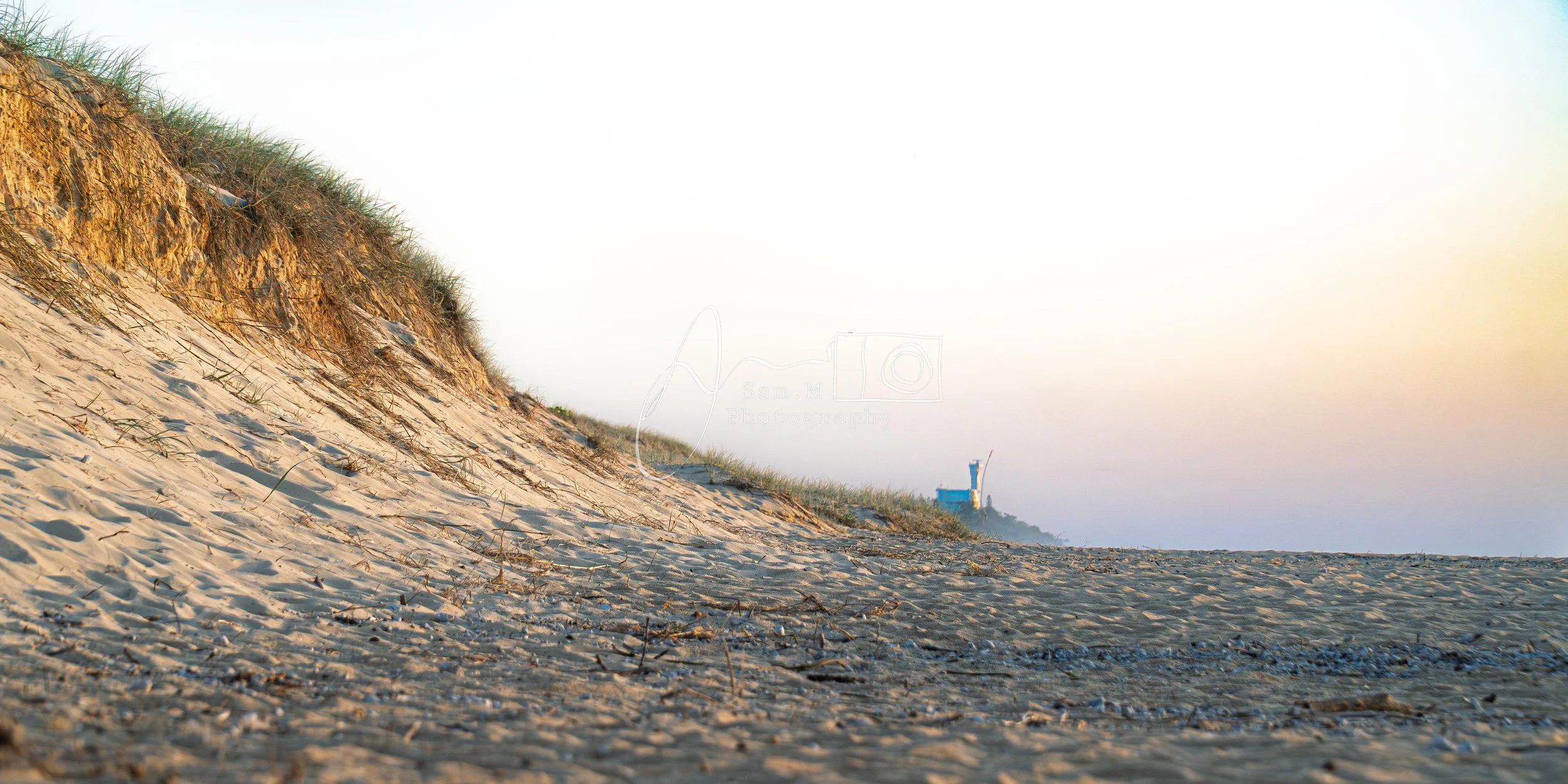 Sandy beach with dunes and grassy vegetation on the left. A distant blue and white lifeguard tower is visible on the right, under a clear sky.