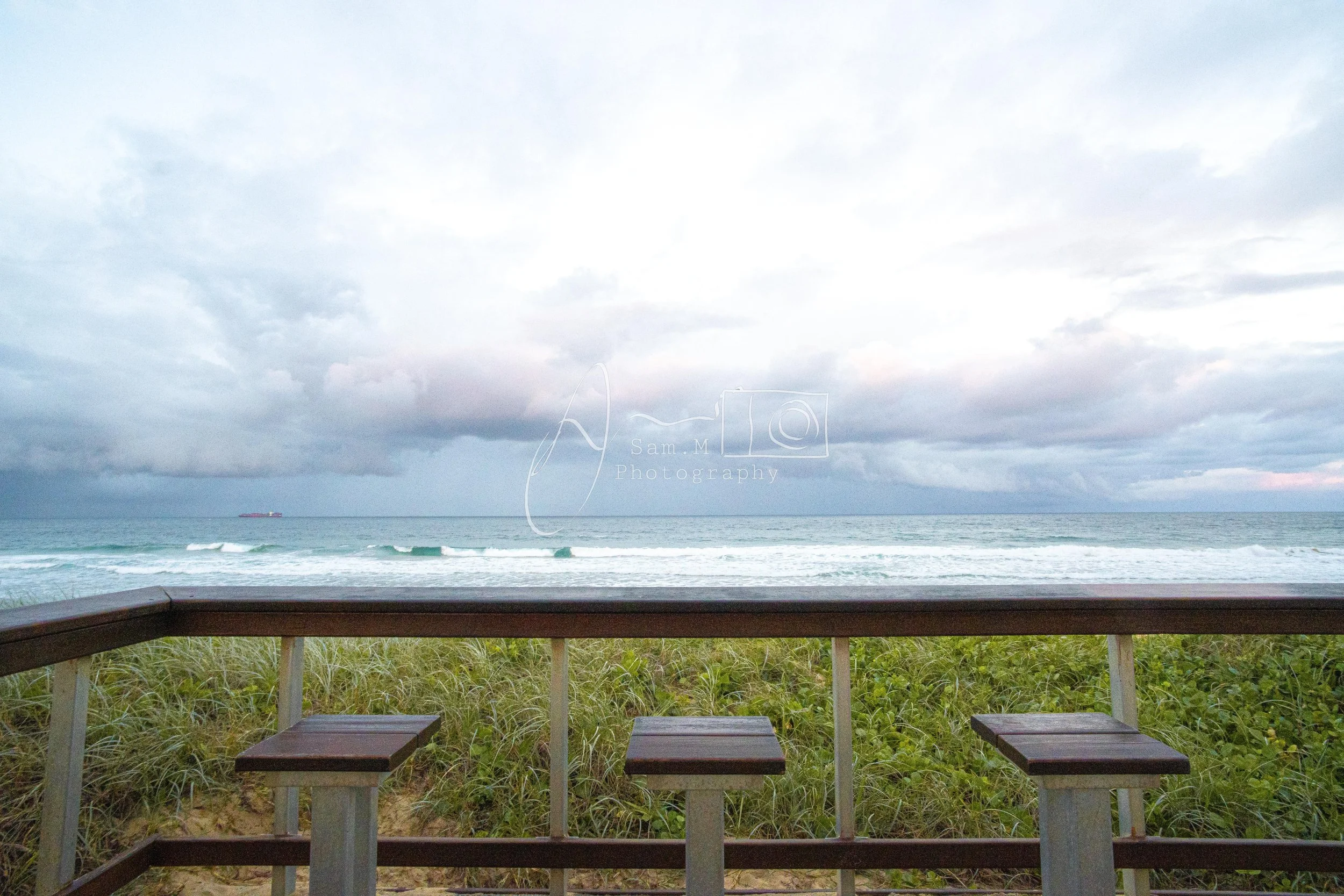 A view of the ocean with gentle waves, lush green dunes, and a wooden seating area with three stools, under a cloudy sky.