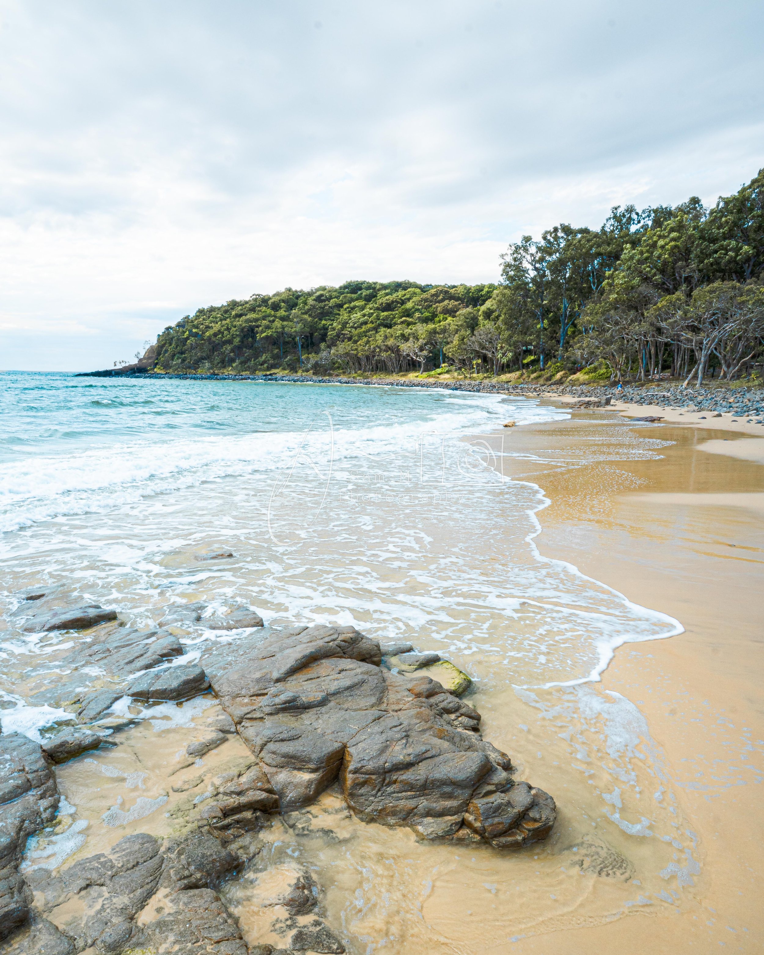 A sandy beach with rocks in the foreground, gentle waves washing onto the shore, a forested hillside in the background, and a cloudy sky overhead.