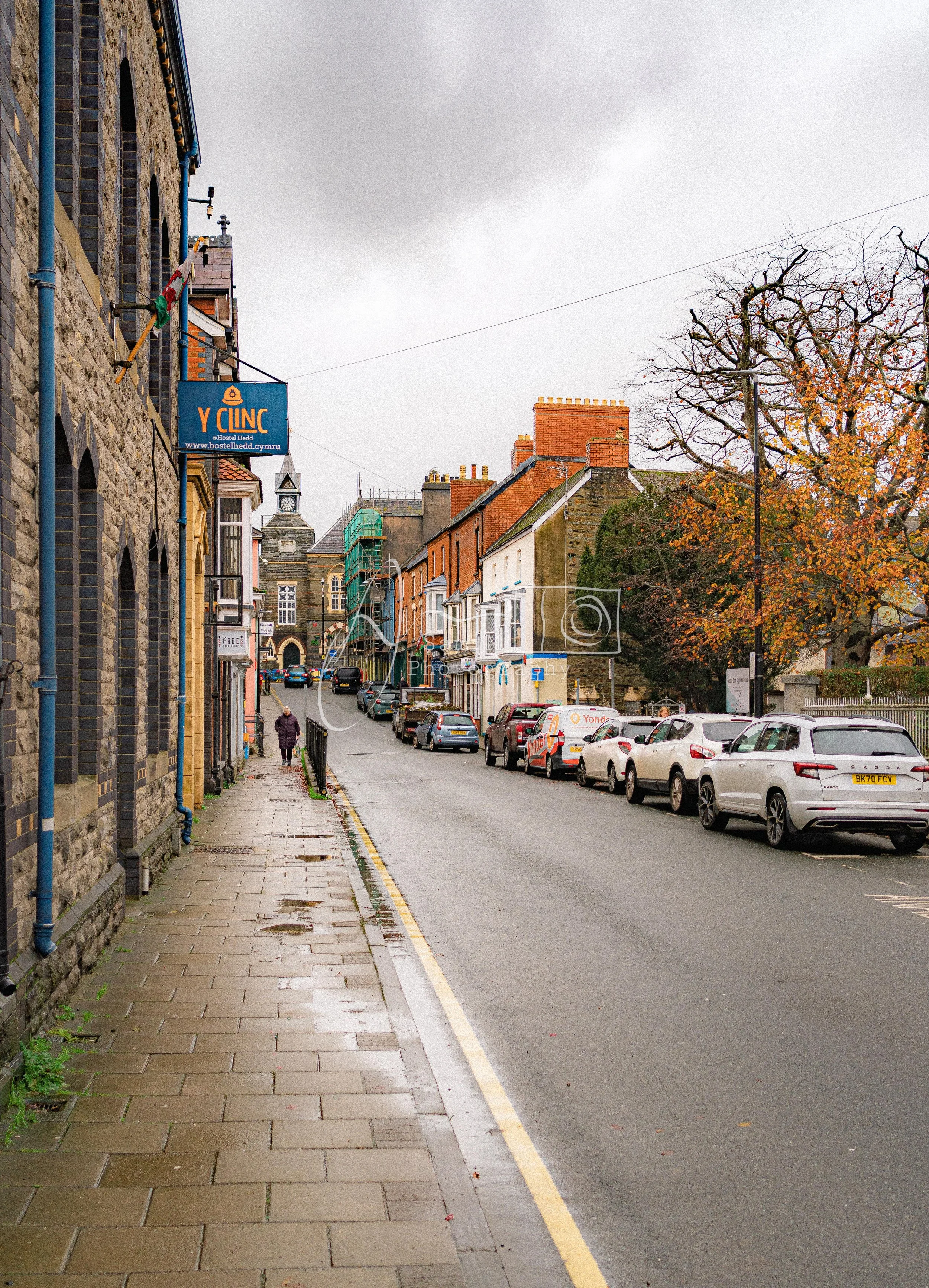 A street scene in a small town with parked cars on the right side, colorful old buildings along the left, and a person walking on the sidewalk. The sky is cloudy, and there are trees with autumn leaves.