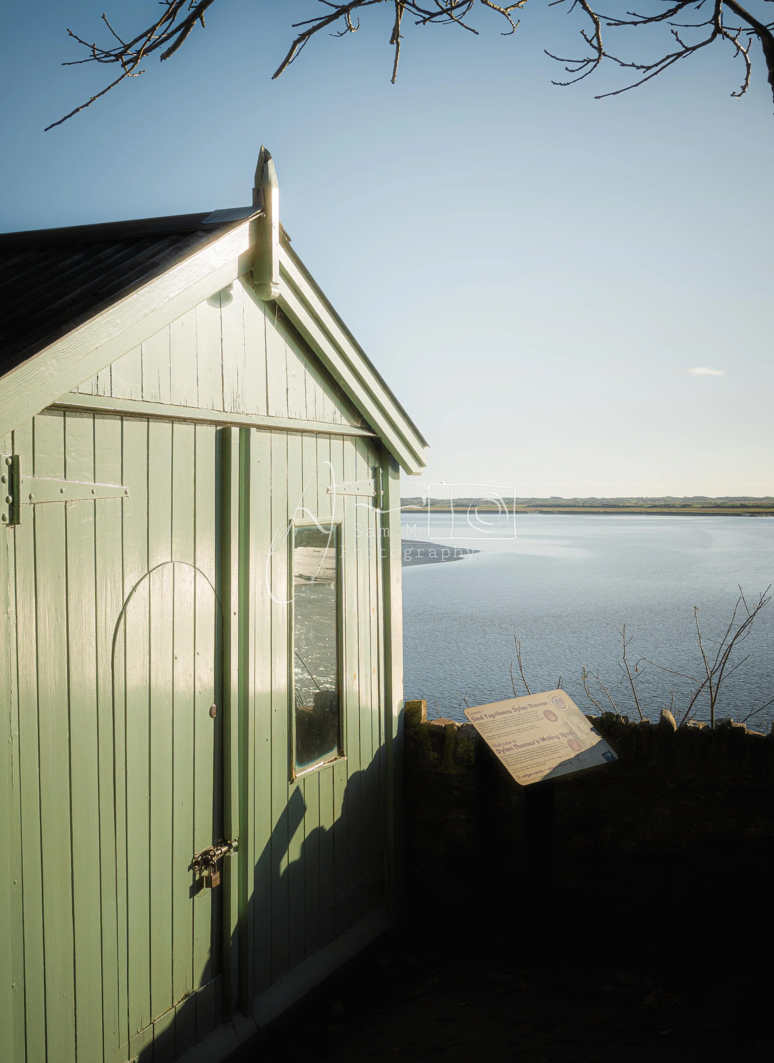 Green wooden shed by a body of water with an information plaque nearby, under a clear sky.