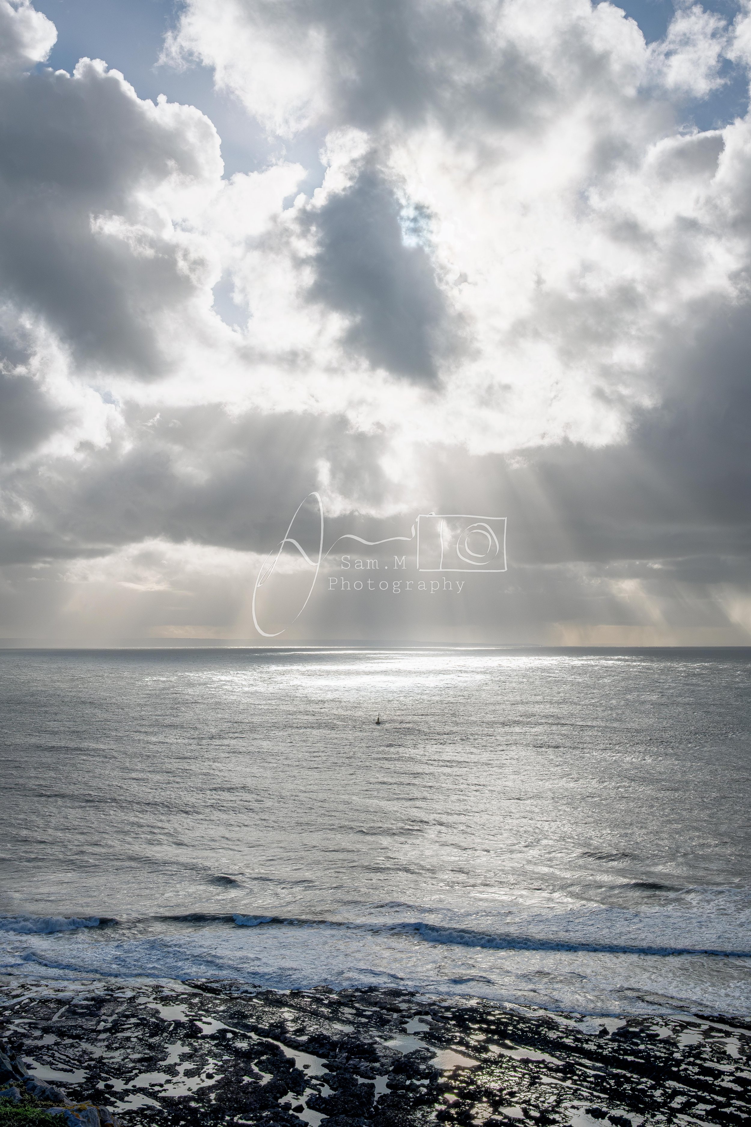 Cloudy sky over ocean with sun rays peeking through clouds, waves crashing on rocks in the foreground.