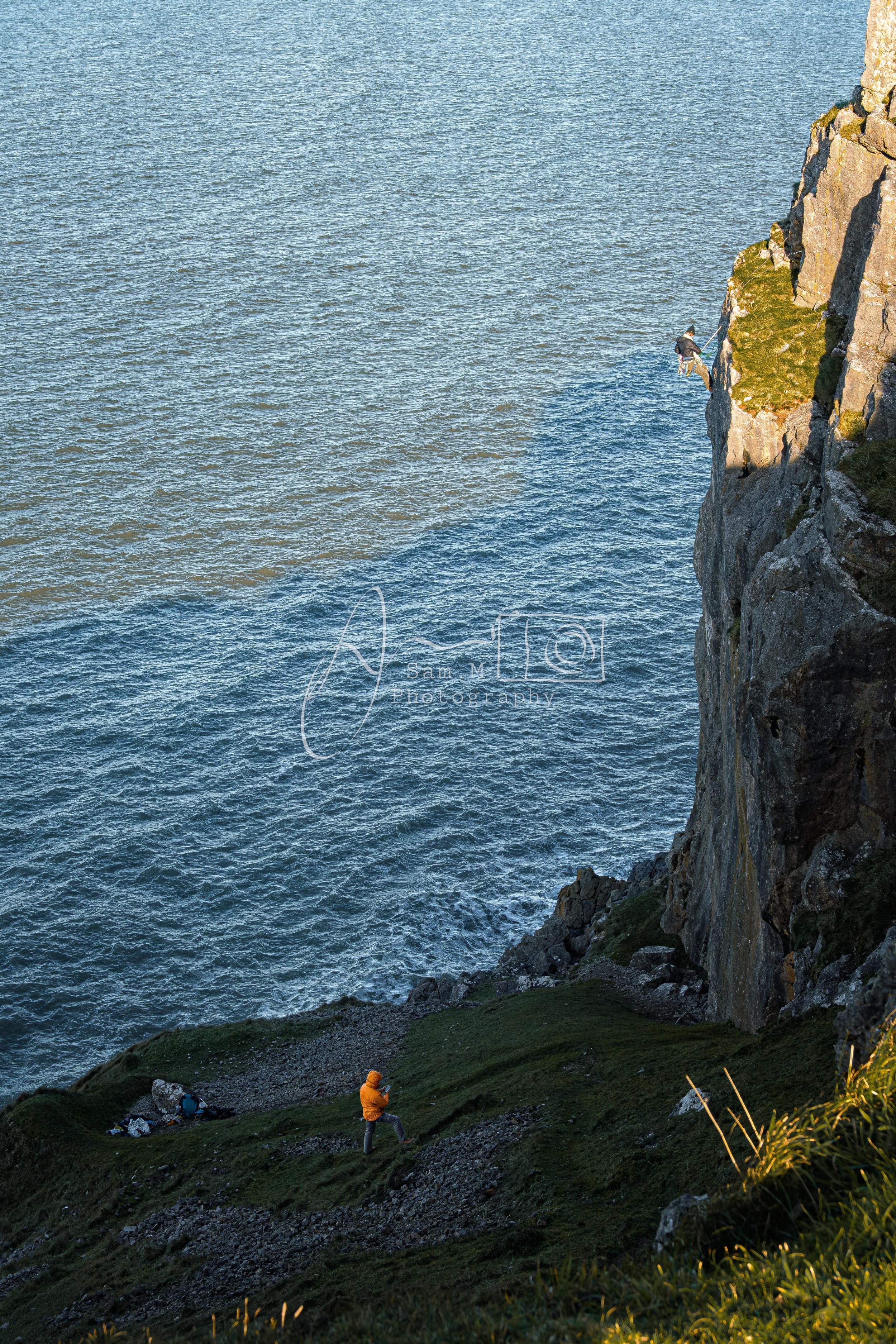 Two people on a grassy cliff overlooking the ocean, one climbing the rocks and the other standing near the edge with a backpack.