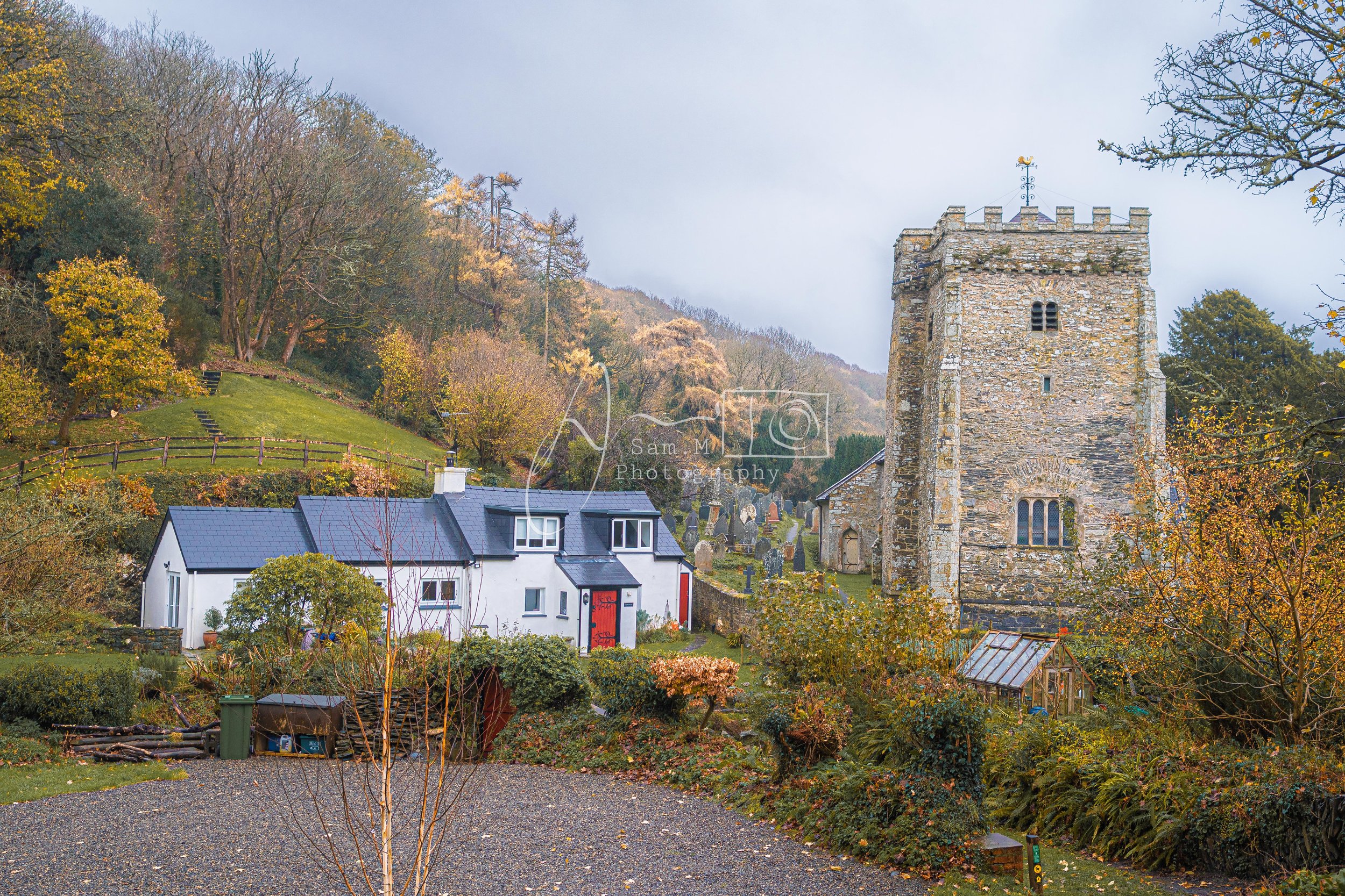 A small white house with a dark roof and a surrounding garden is situated at the base of a hill. Behind it, an ancient stone church with a square tower is visible. The hill is covered with trees and grass, with some autumn leaves, and there are grave