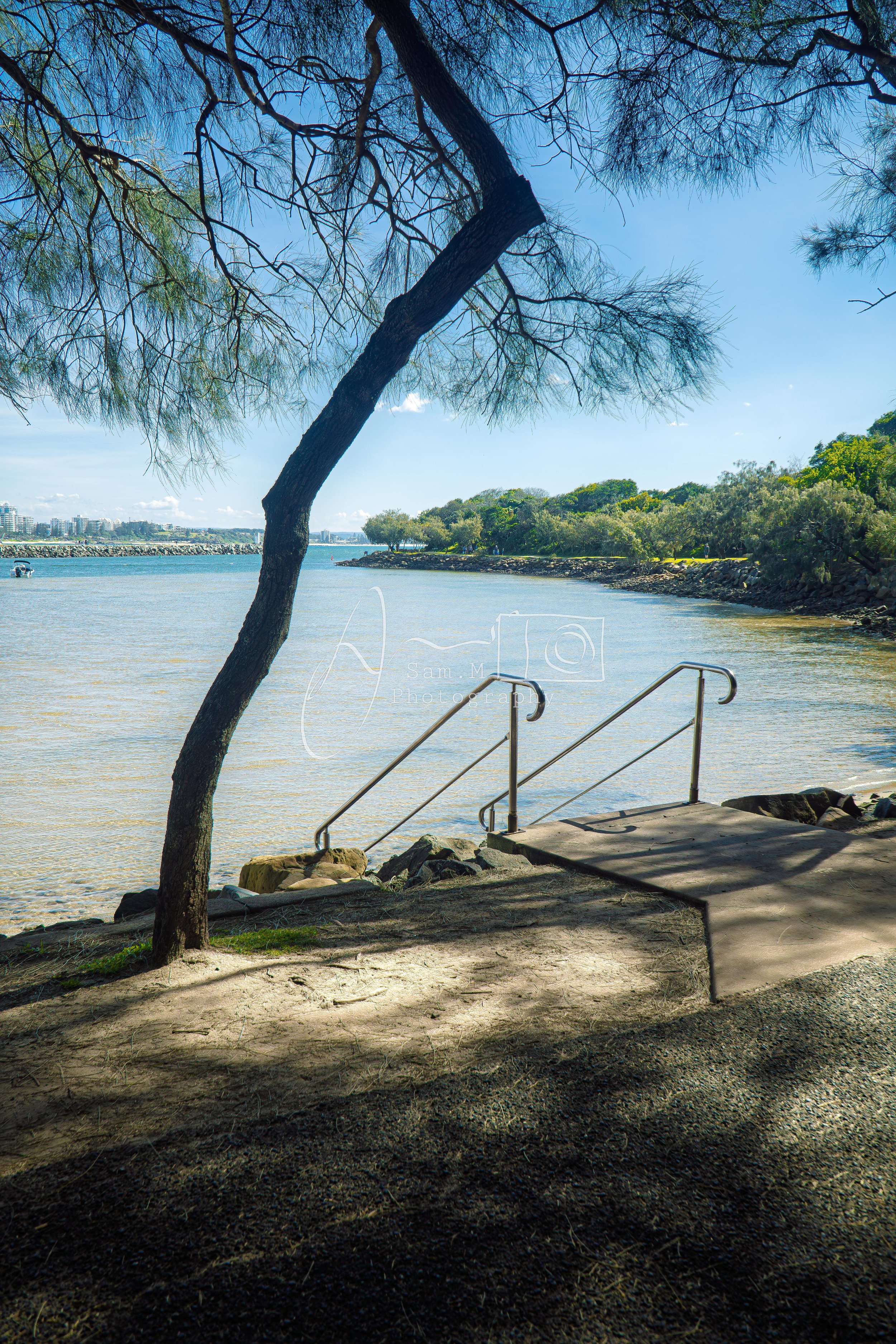 A scenic view of a river with a concrete boat ramp and metal handrails leading into the water. A large tree with sprawling branches provides shade over the ramp, and the background features green trees and a clear blue sky.