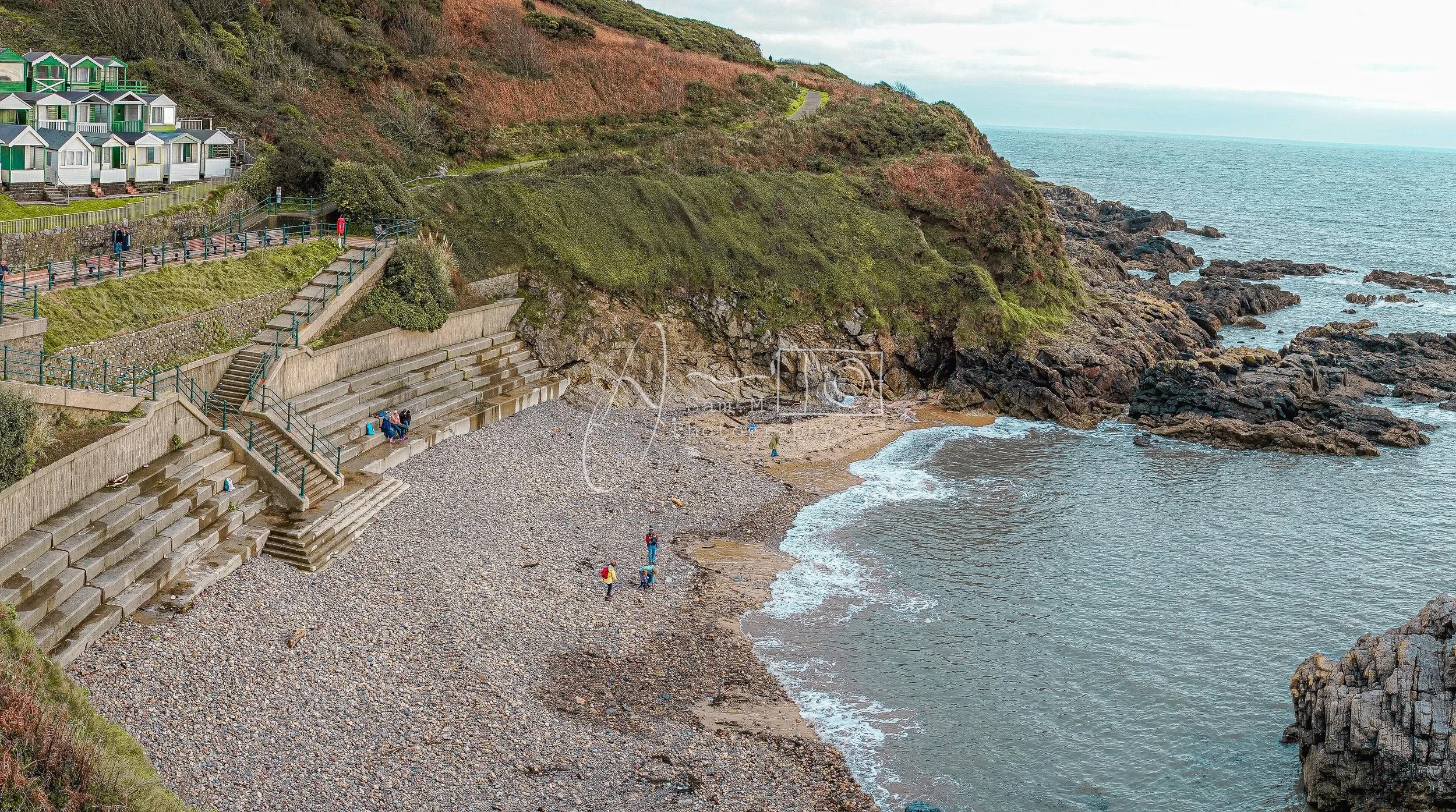 People walking and sitting on concrete steps at a pebble beach with rocky cliffs and ocean in the background, overlooking a water inlet with waves gently hitting the shore.