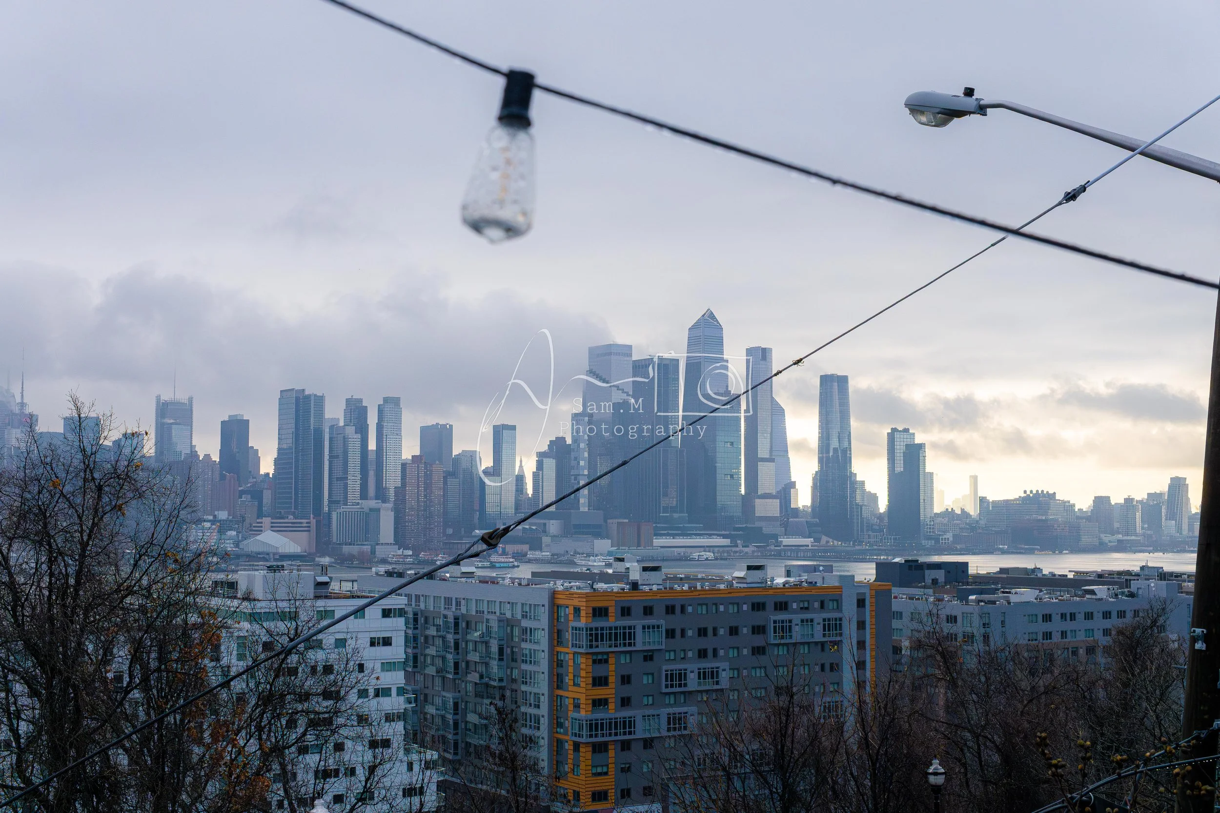 City skyline with tall skyscrapers viewed through overhead string lights and power lines, overcast sky with clouds.