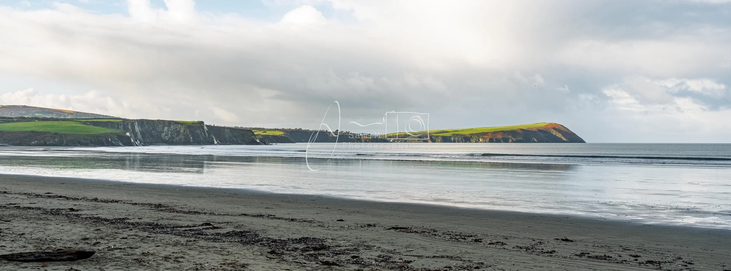 Scenic view of a beach with dark sand, calm water, and cliffs with green grass and rocky outcroppings under a cloudy sky.