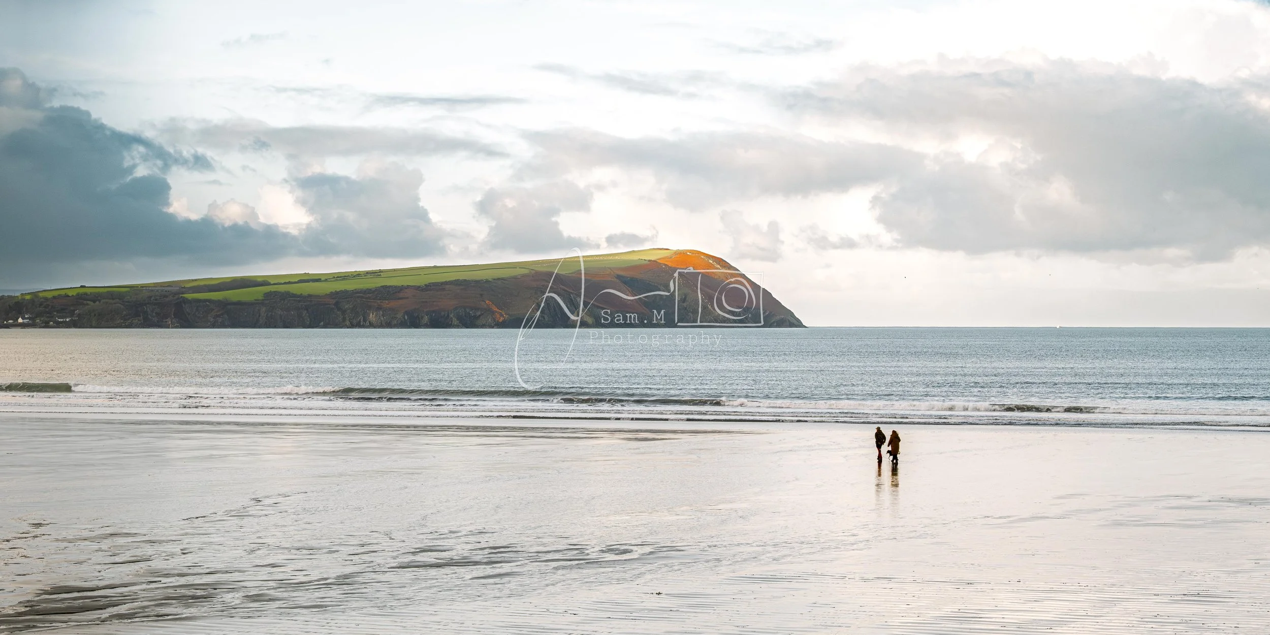 Two people walking on the wet beach shoreline with the ocean and a distant green cliff in the background under a cloudy sky.