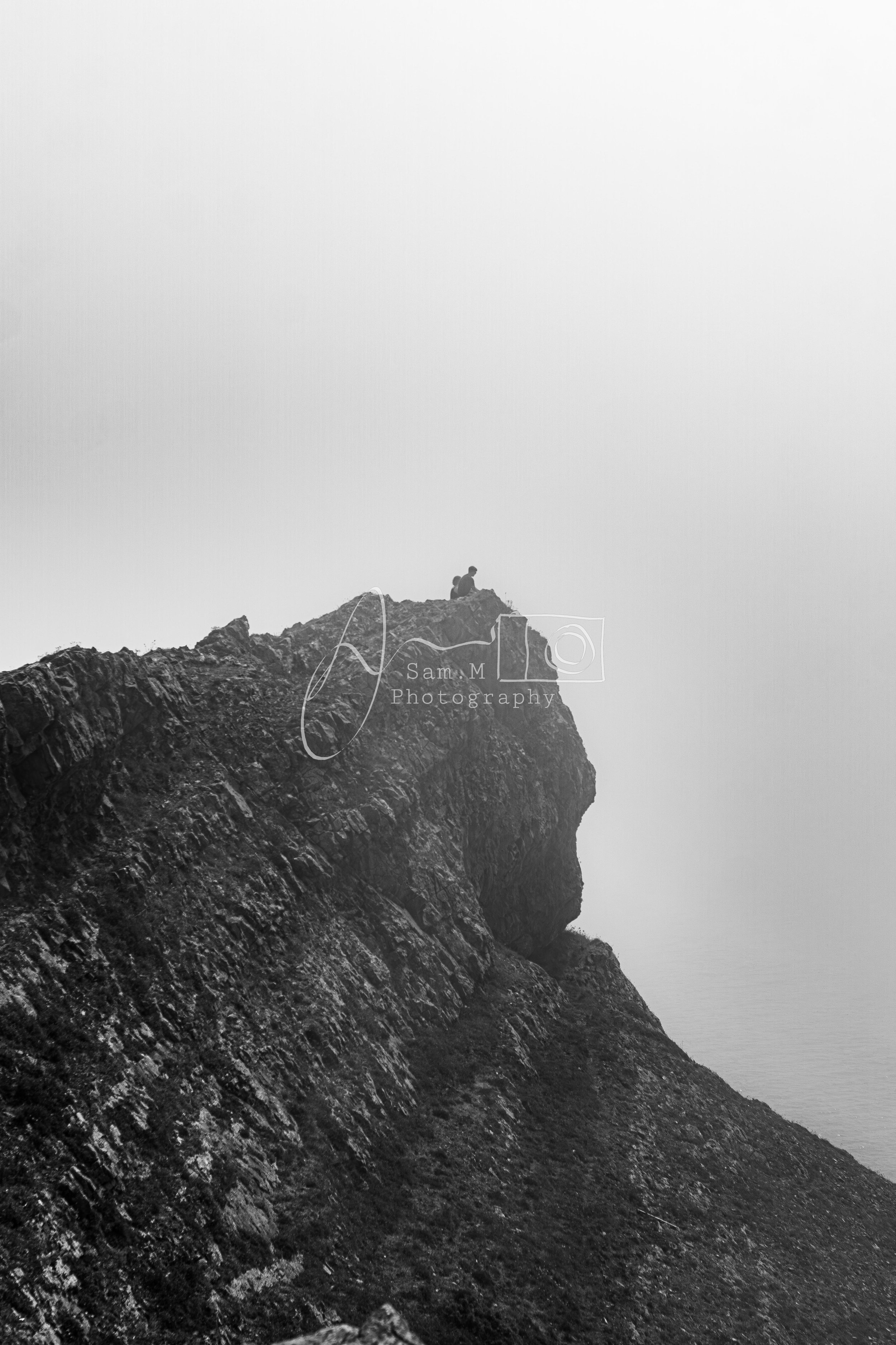 A person sitting on top of a rocky mountain peak, overlooking the foggy landscape below, in black and white.