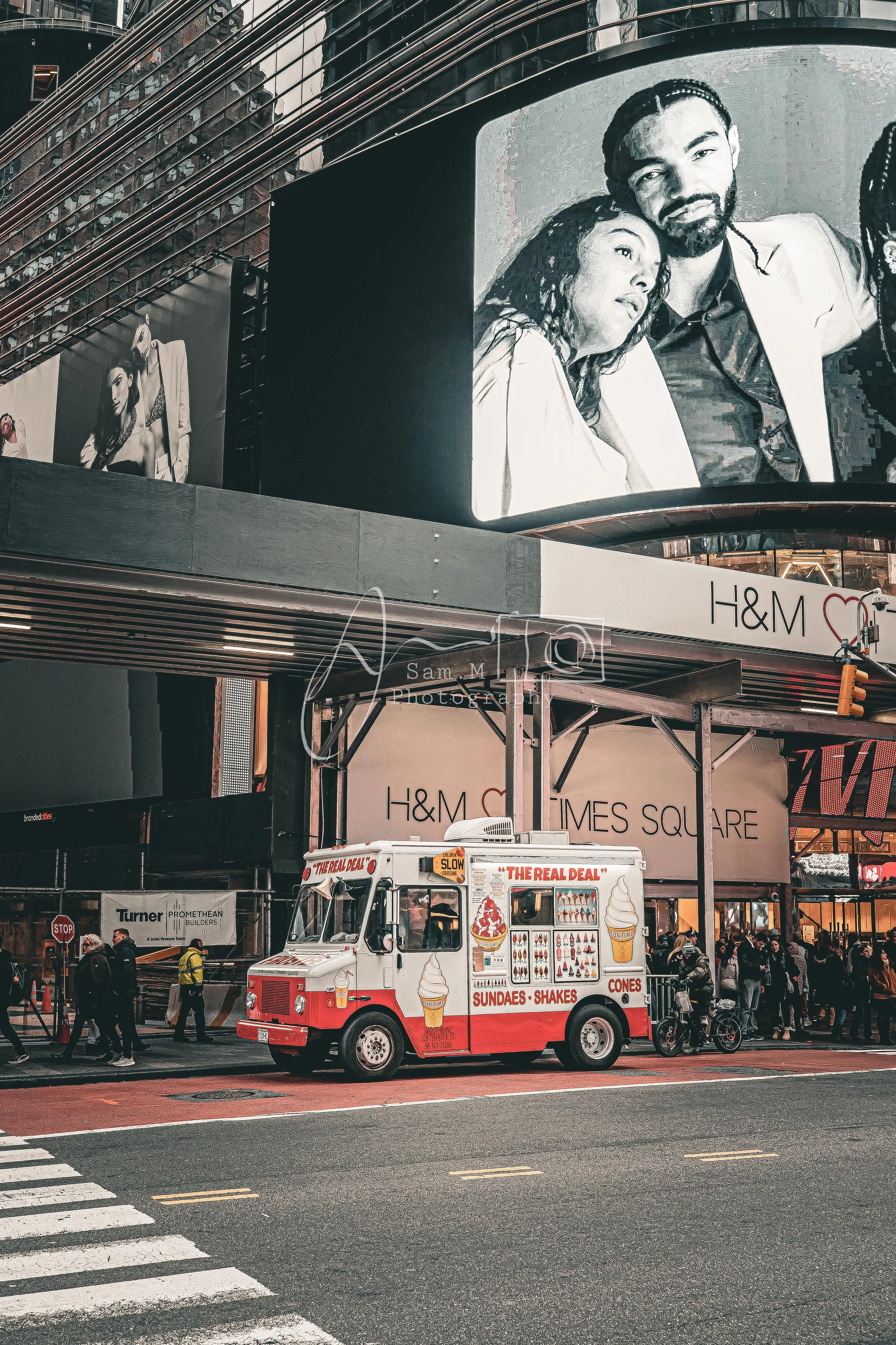 A street scene featuring a red and white ice cream and dessert truck, a crowd of people, and billboards including one with two people and another with a fashion model, in Times Square, New York City.
