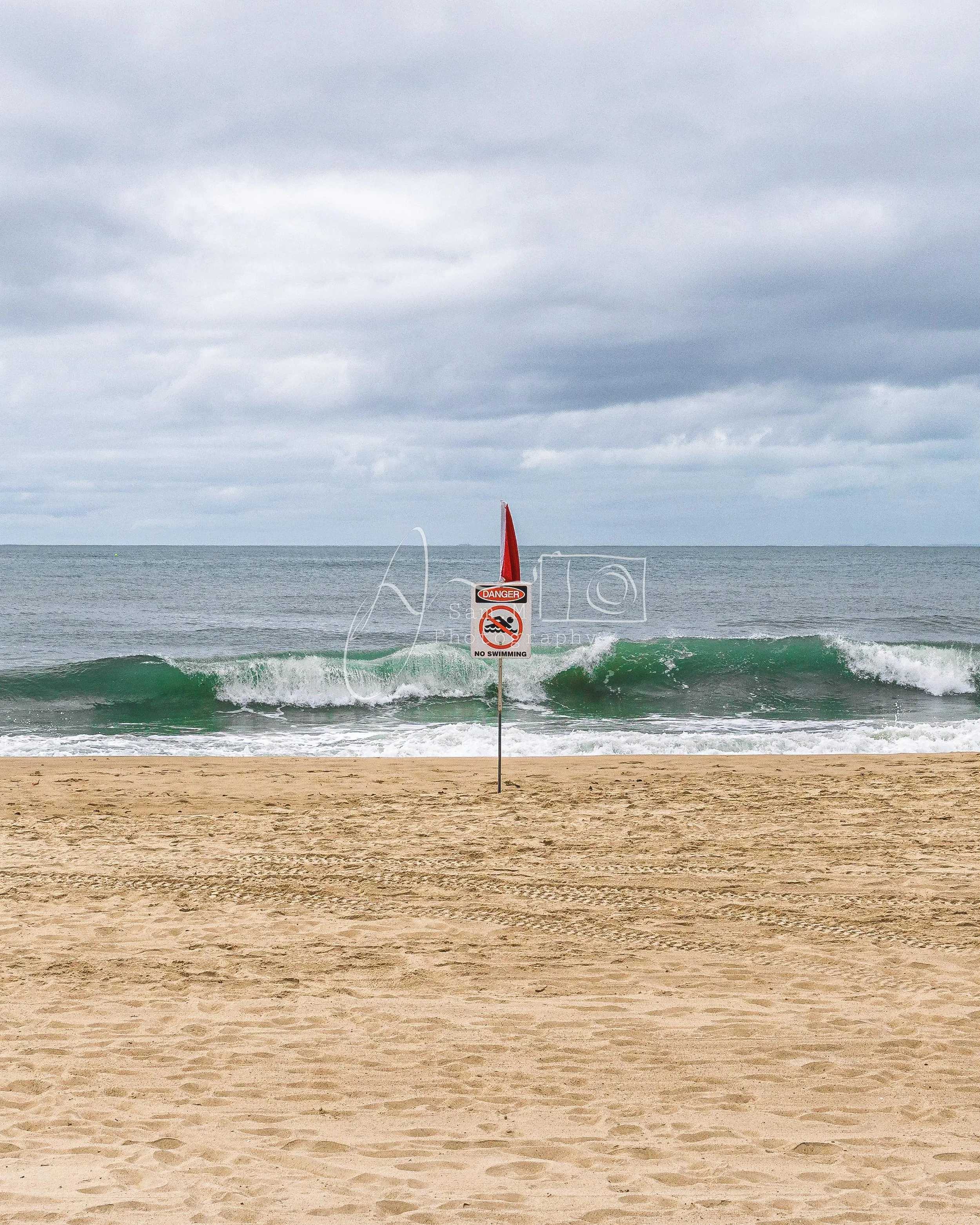 A sandy beach with a red warning flag and a 'No Swimming' sign planted in the sand, with ocean waves and a cloudy sky in the background.