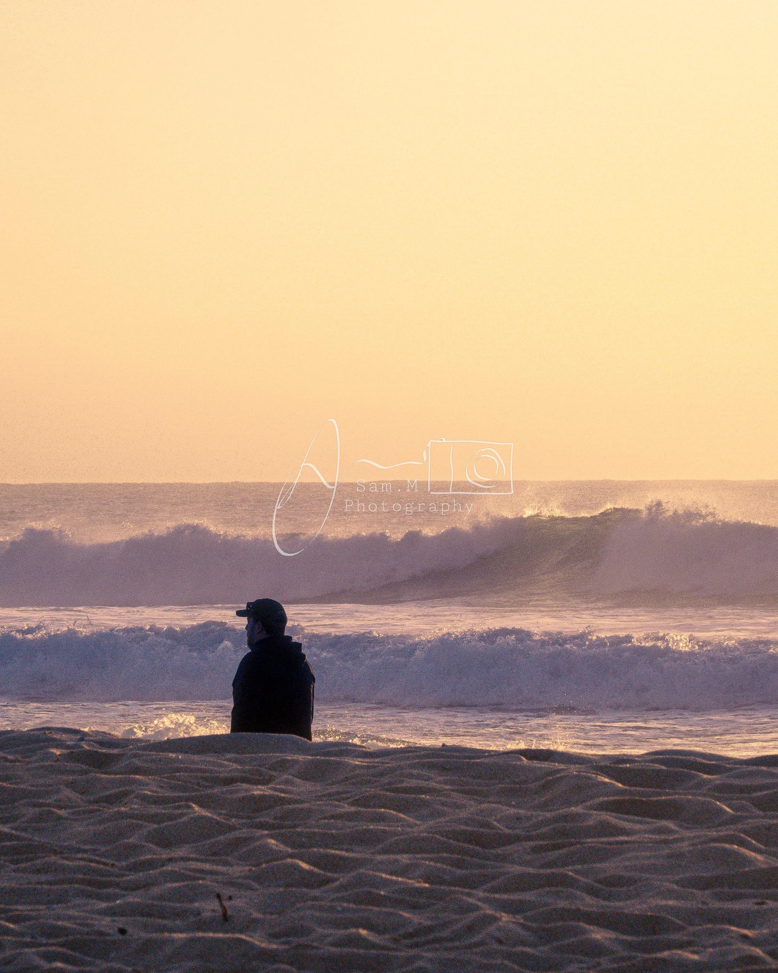 A person standing on the sandy beach, looking out at the ocean waves during sunset.