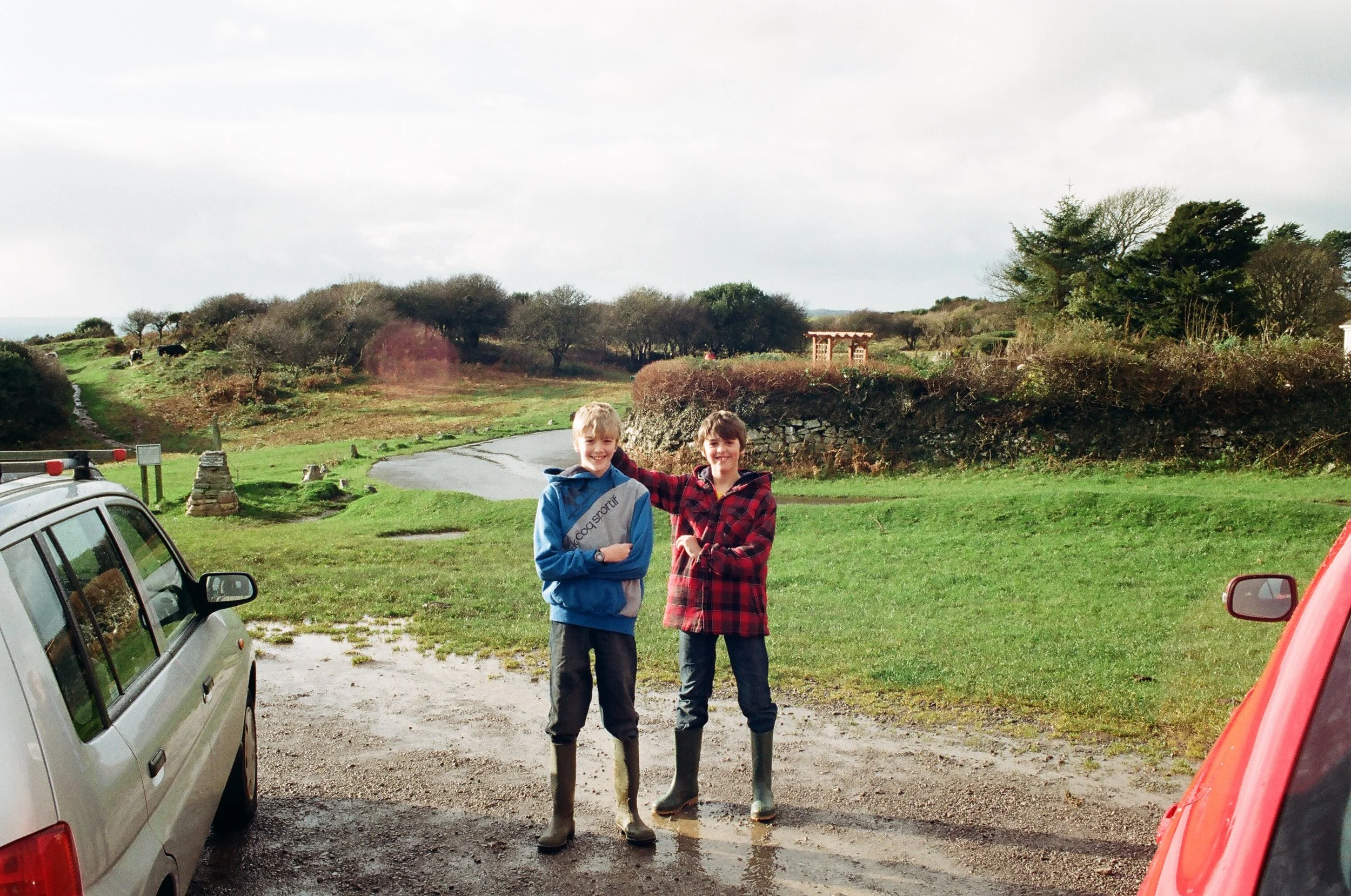Two children standing outdoors on a gravel driveway near parked cars with a grassy field, trees, and cloudy sky in the background.