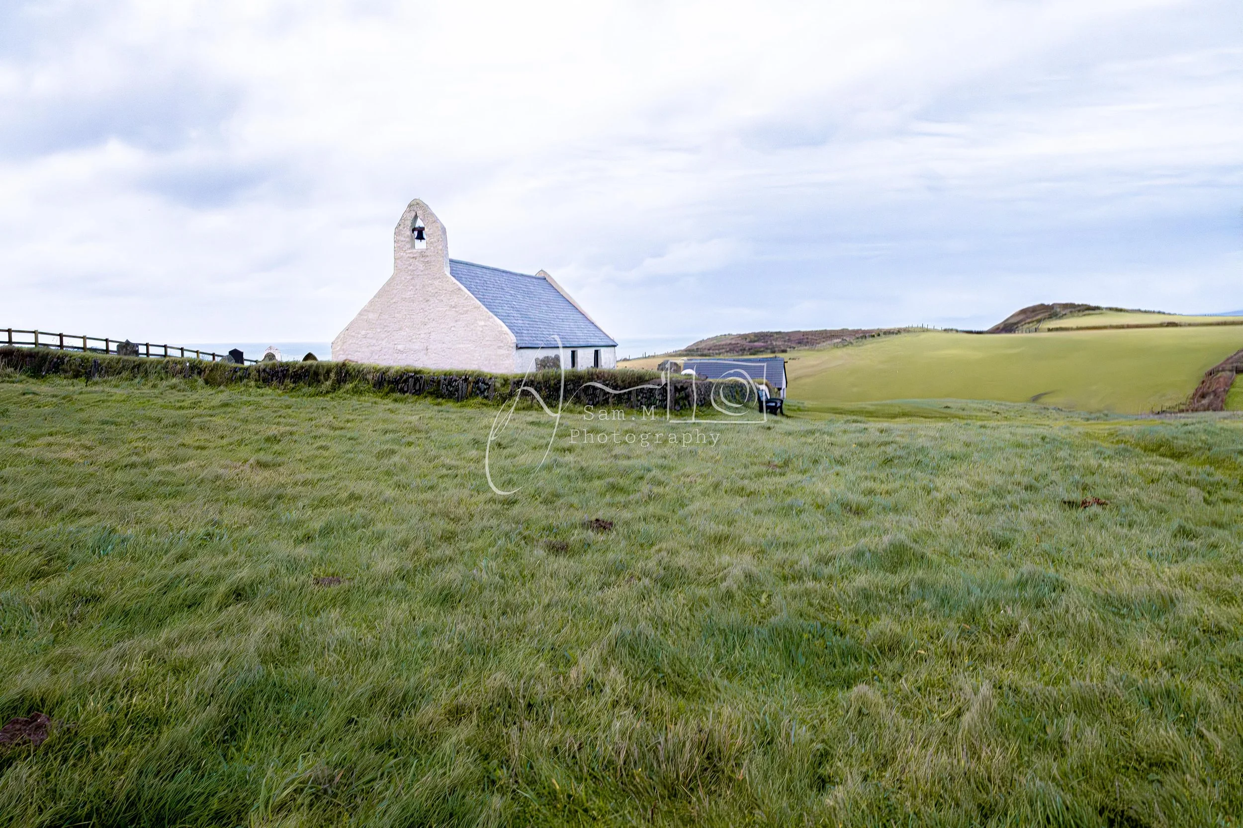 A small white stone church with a bell tower is situated on a grassy hillside, with rolling hills in the background under a cloudy sky.