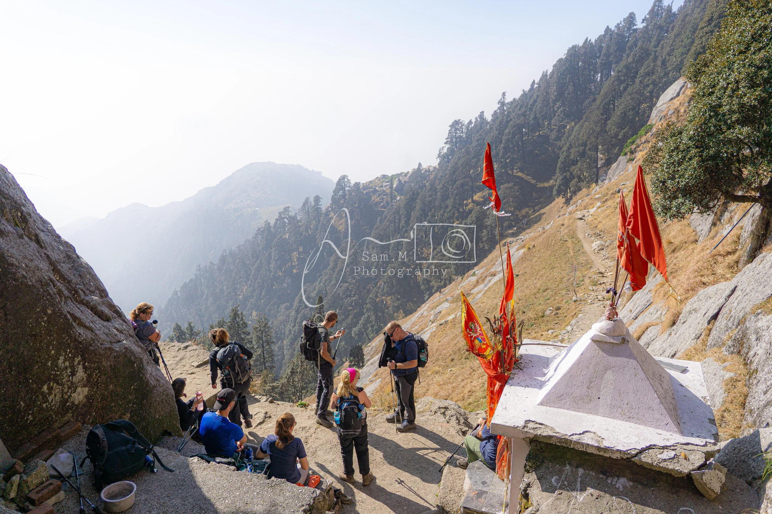 Group of hikers with backpacks gathered around a small shrine with red flags, situated on a mountain trail with forested slopes and distant mountains in the background.