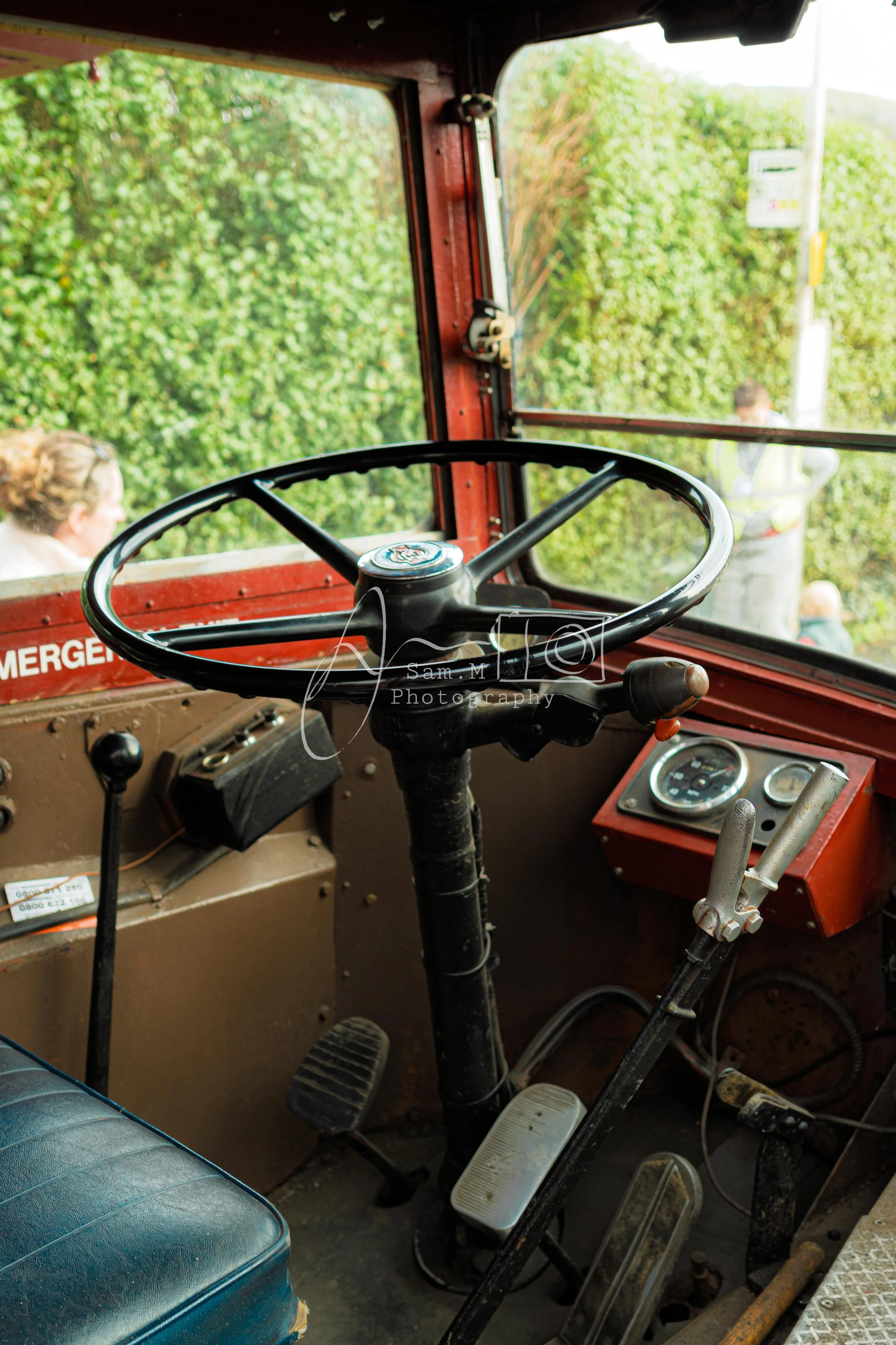 Interior of an old industrial or emergency vehicle cab, featuring a large black steering wheel, pedals, and various gauges and controls, with a window showing greenery and a person outside.