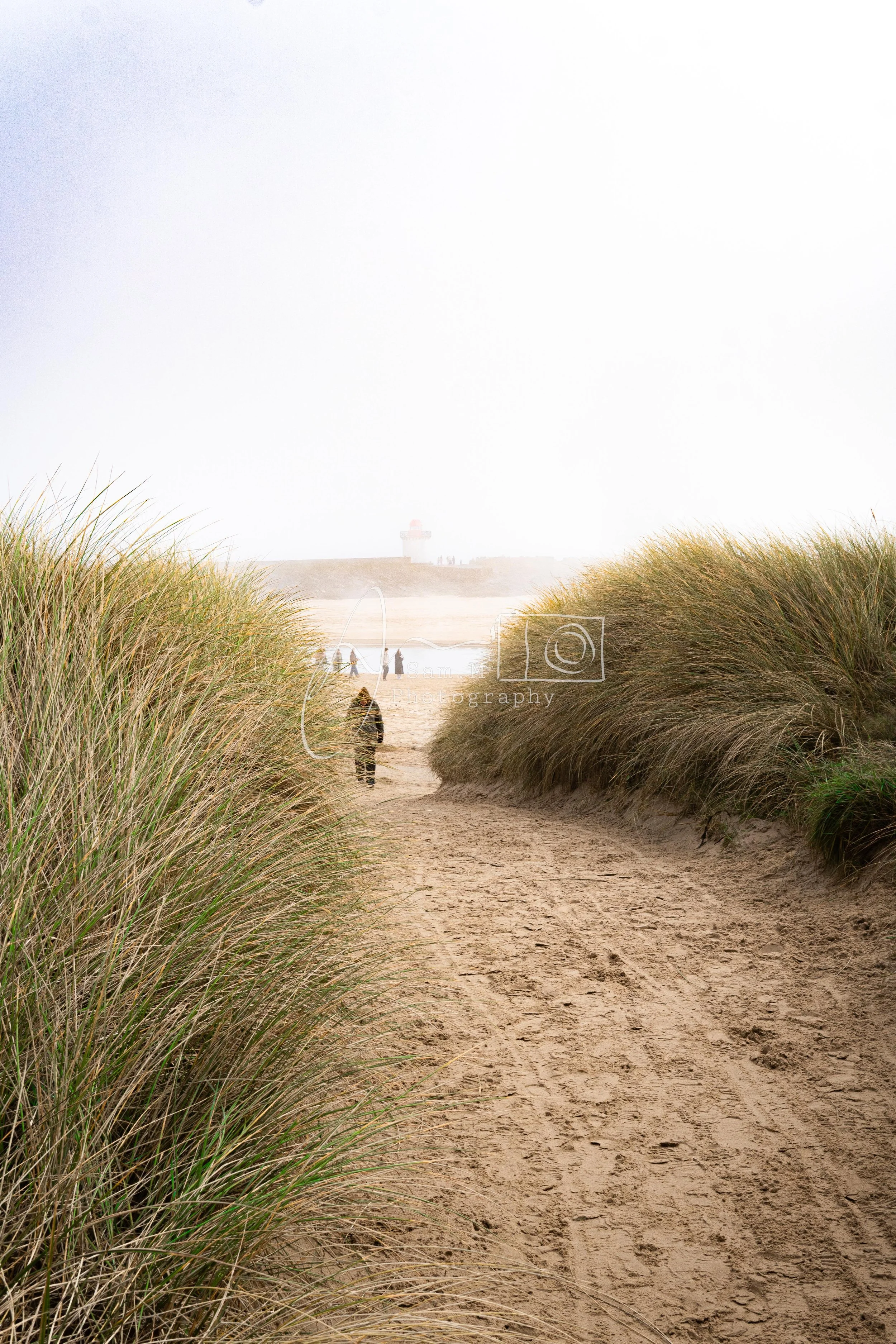 A sandy pathway leading through tall grasses towards a beach, with a few people walking and a lighthouse in the background.