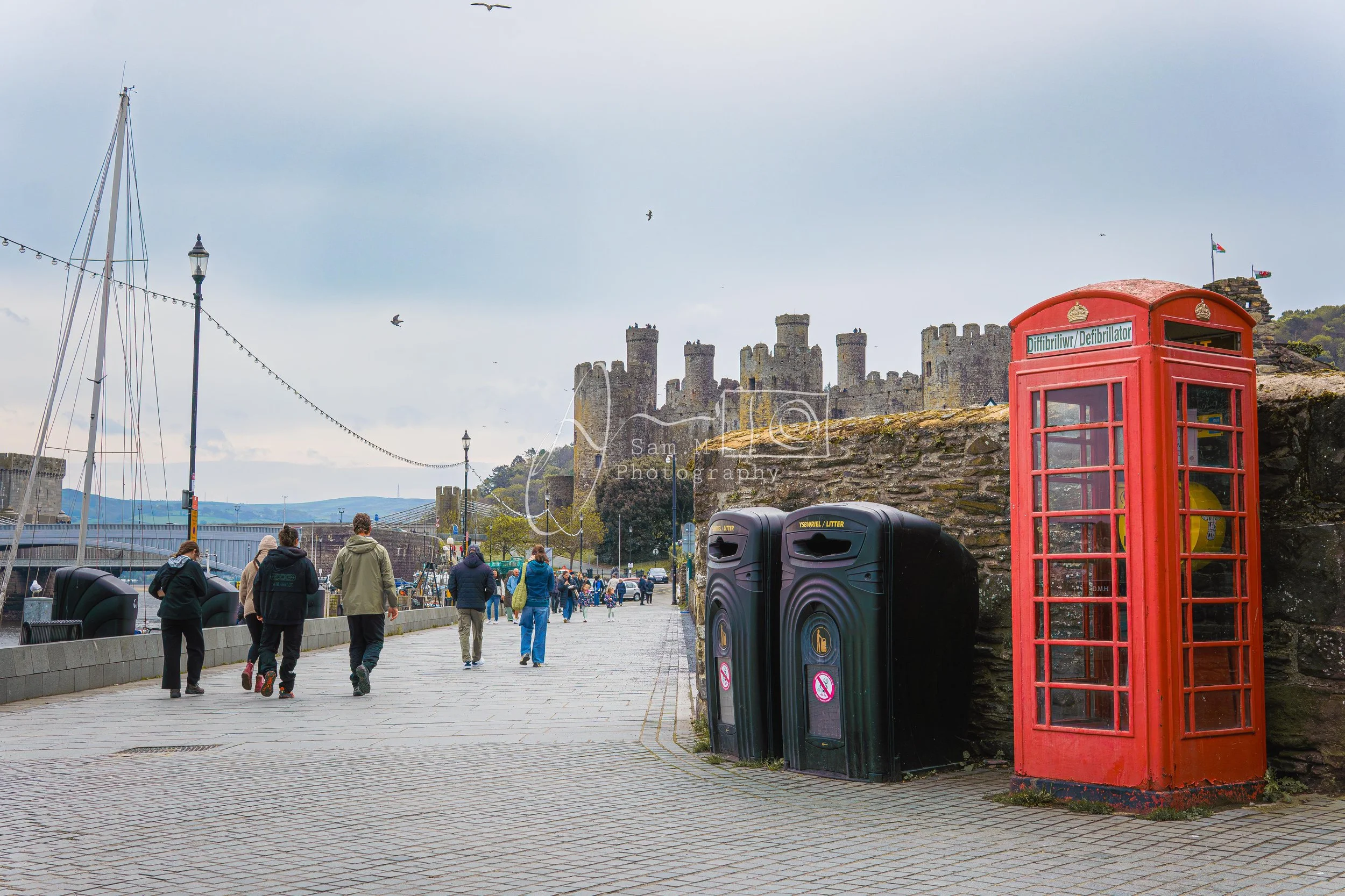 A harbor promenade with people walking, a traditional red telephone booth, black trash bins, historic castle on a hill, sailboats docked nearby, and a cloudy sky with birds flying.