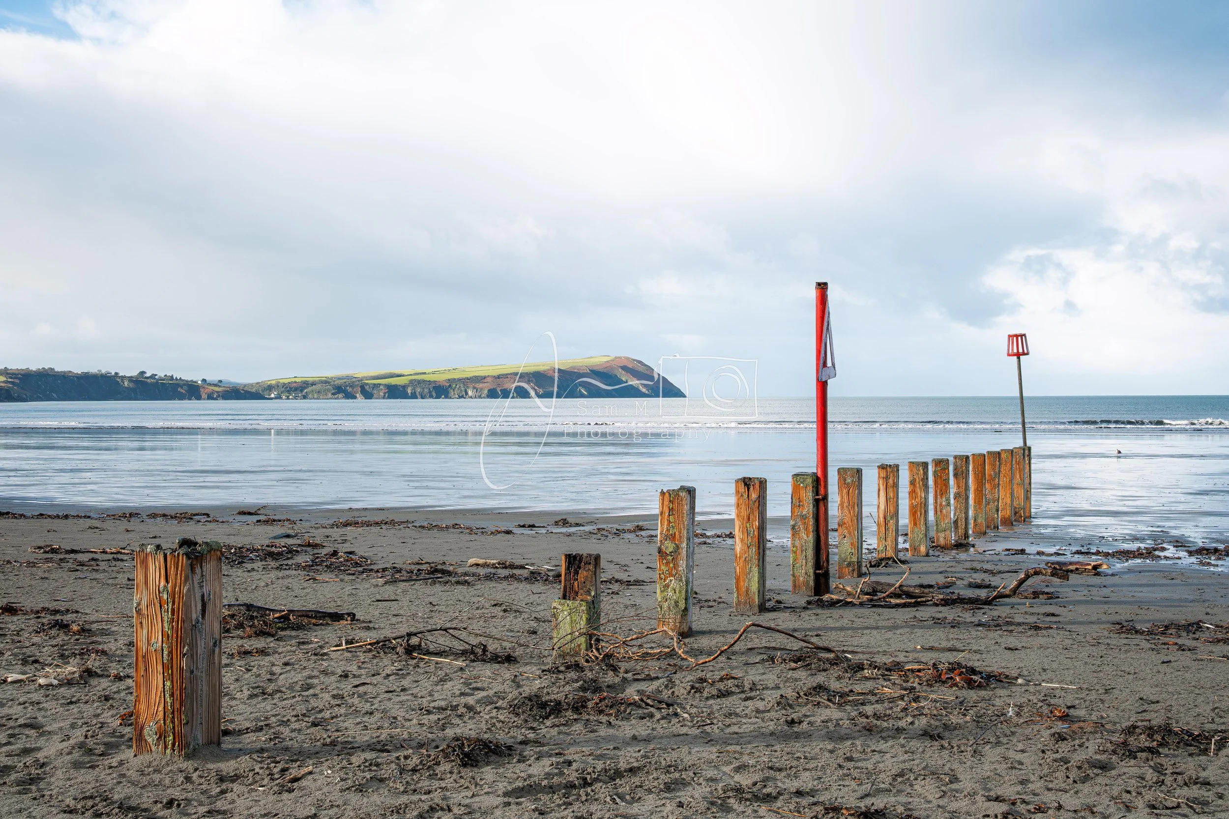 Empty beach with wooden posts, red rescue signs, and a distant landmass under cloudy sky.