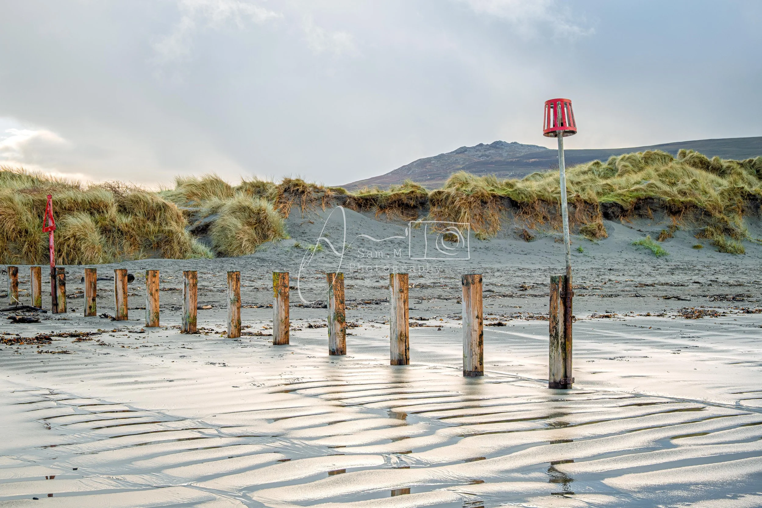 Seaweed-covered sand dunes with wooden posts and red fishing shelters on a beach, with grassy dunes and mountains in the background.