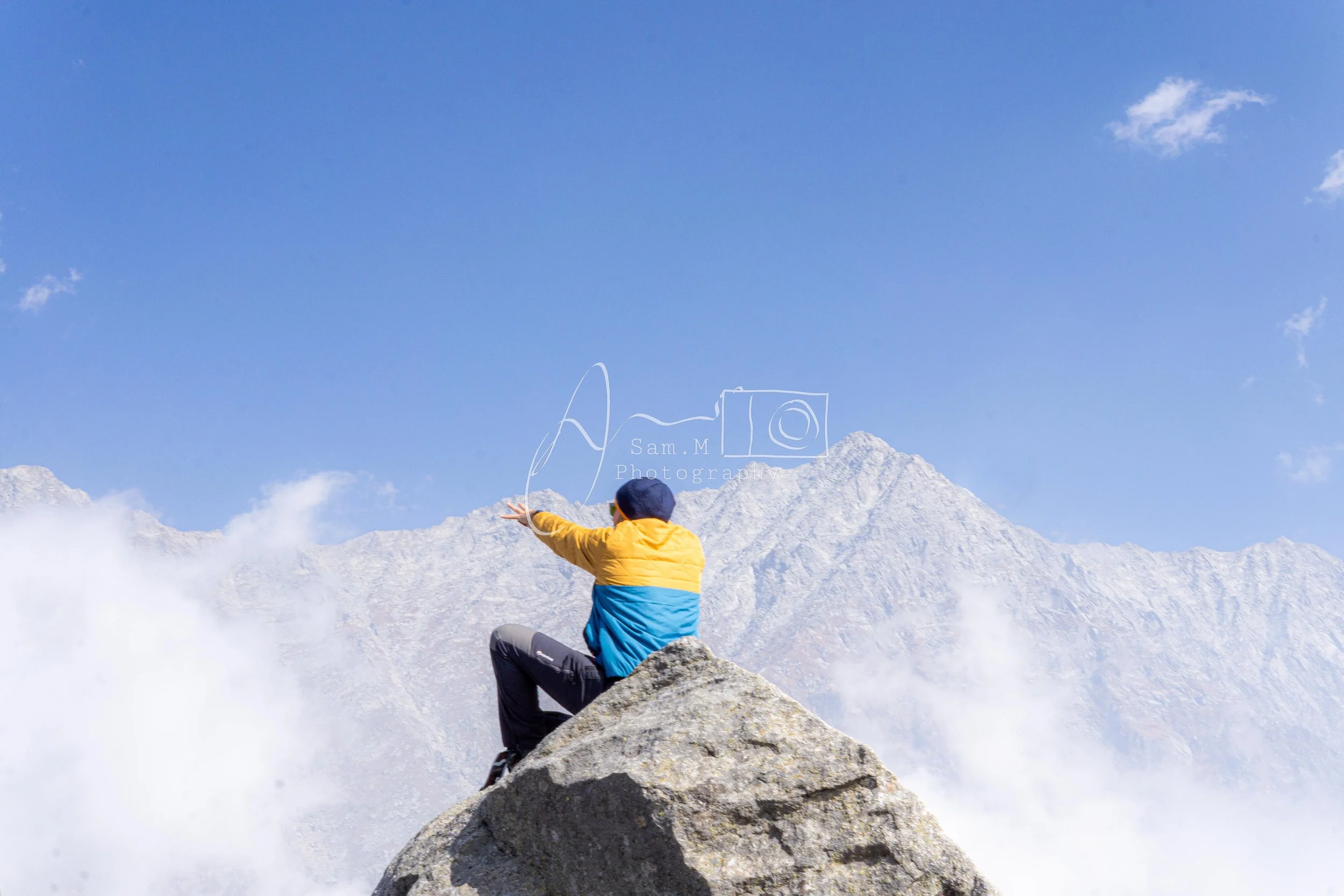 Person sitting on a large rock on a mountain peak, facing away, wearing a yellow and blue jacket, with a mountain range and blue sky in the background.