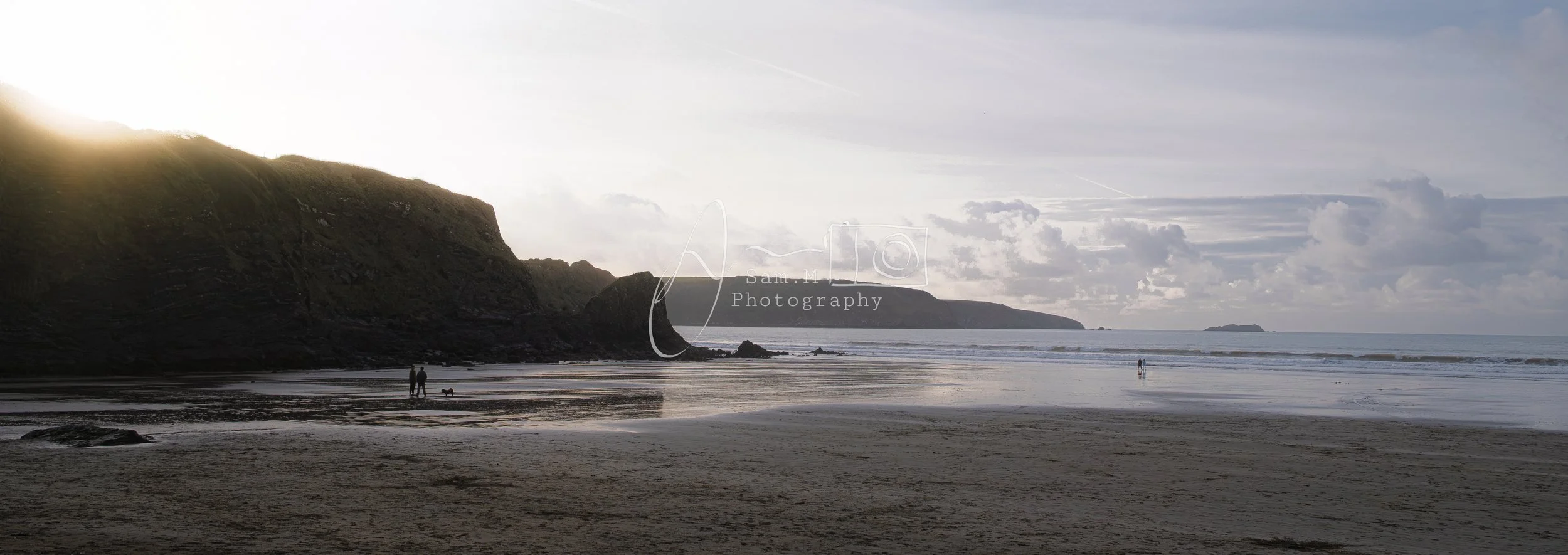 A beach scene at sunset with cliffs on the left, the ocean on the right, and a few people walking along the shore.