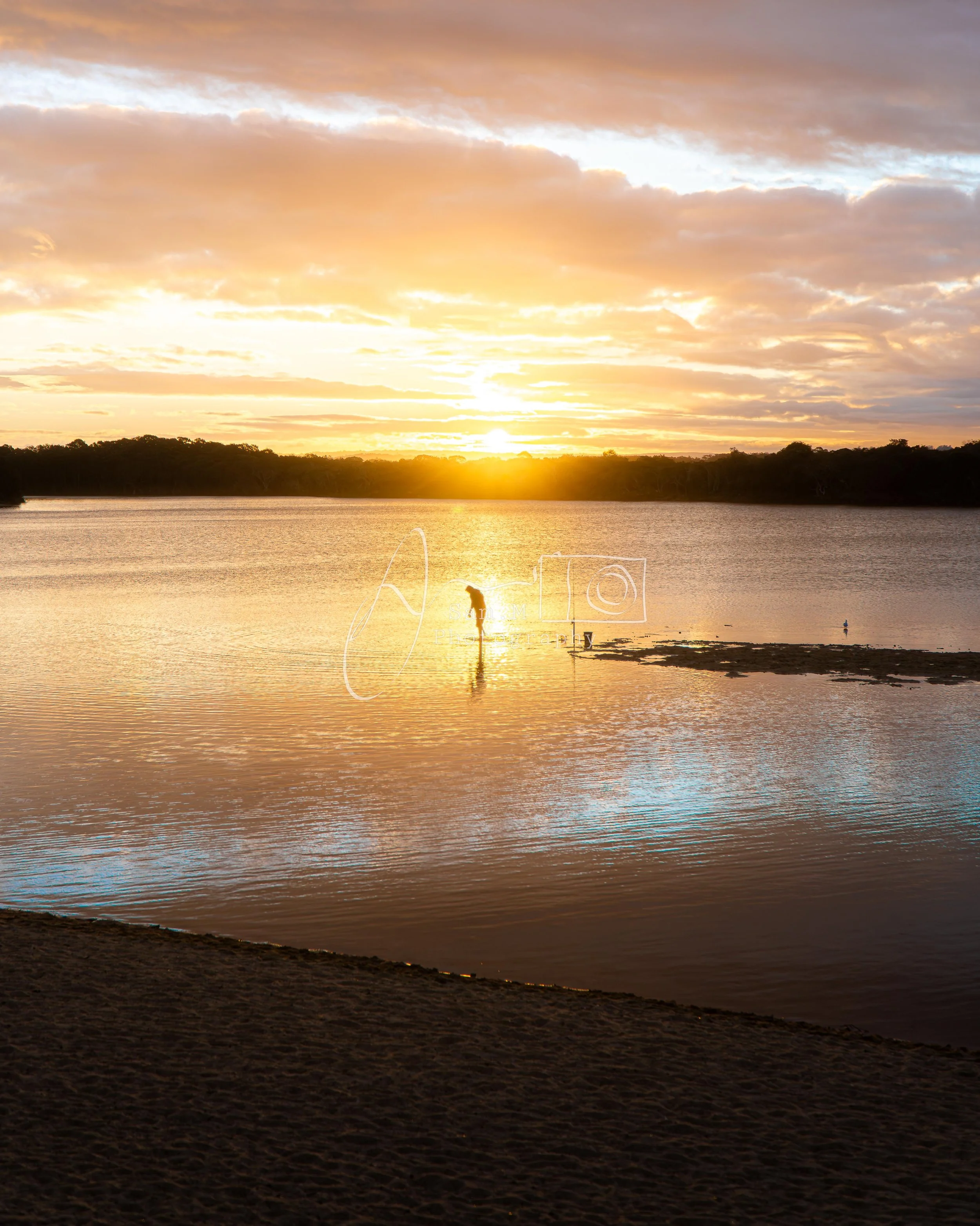 A person stands in shallow water at sunset, with the sky filled with orange and purple clouds, and trees lining the horizon.