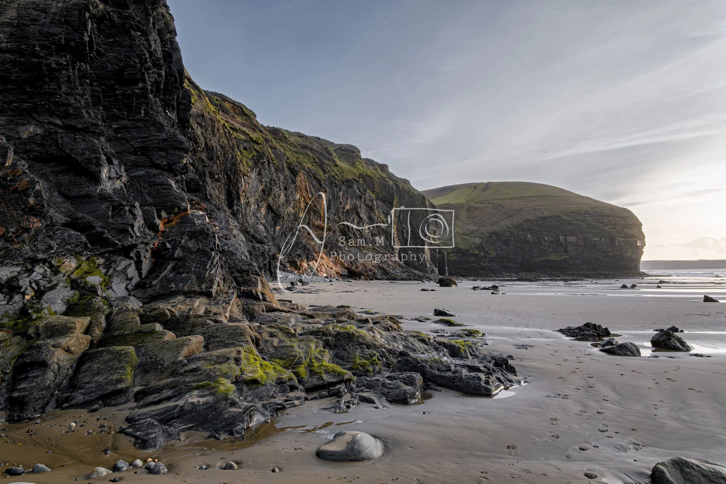 Scenic view of a beach with rocky cliffs on the left and the ocean on the right, under a partly cloudy sky.