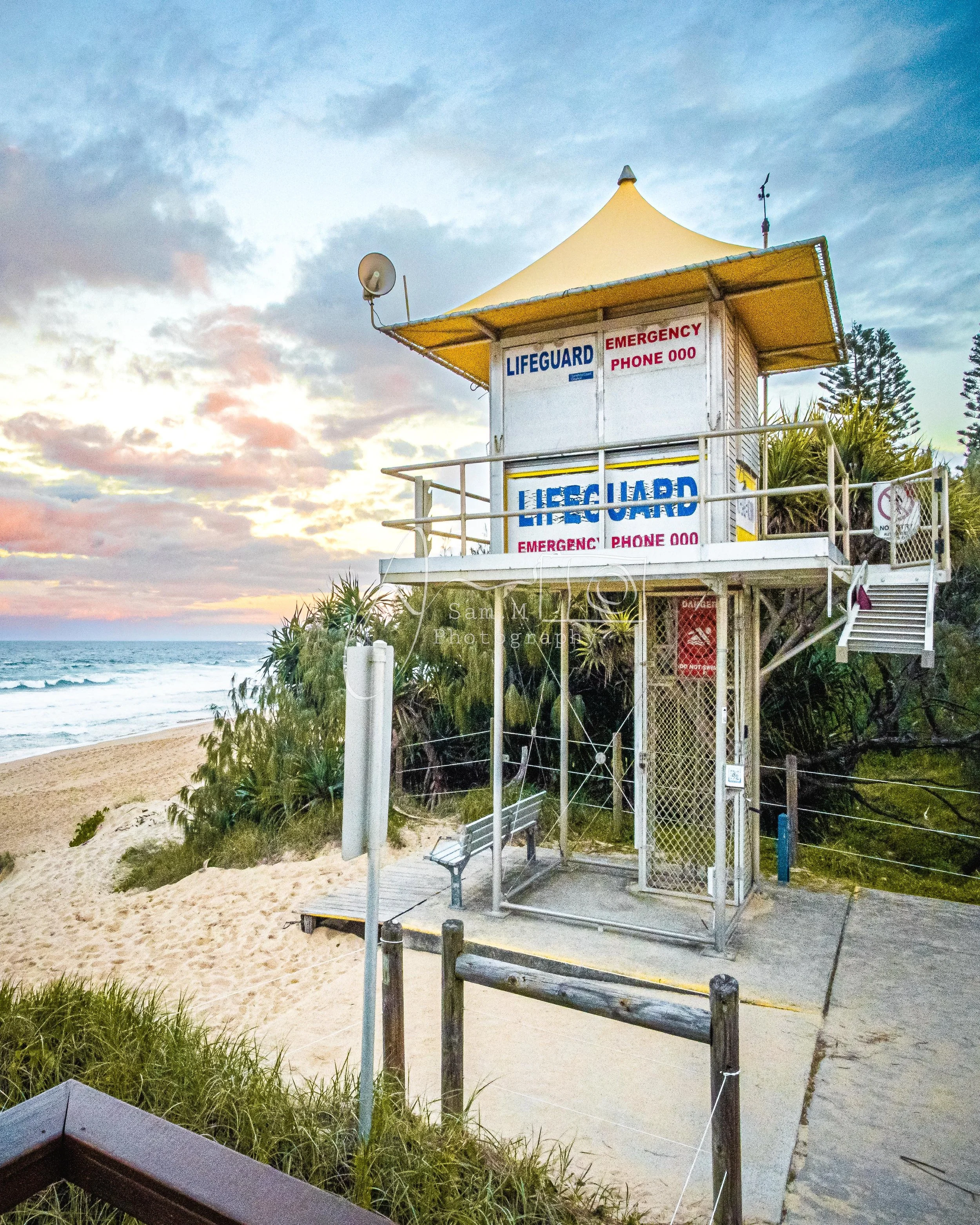 A lifeguard tower on a beach at sunset with a sandy shore, ocean waves, and cloudy sky in the background.