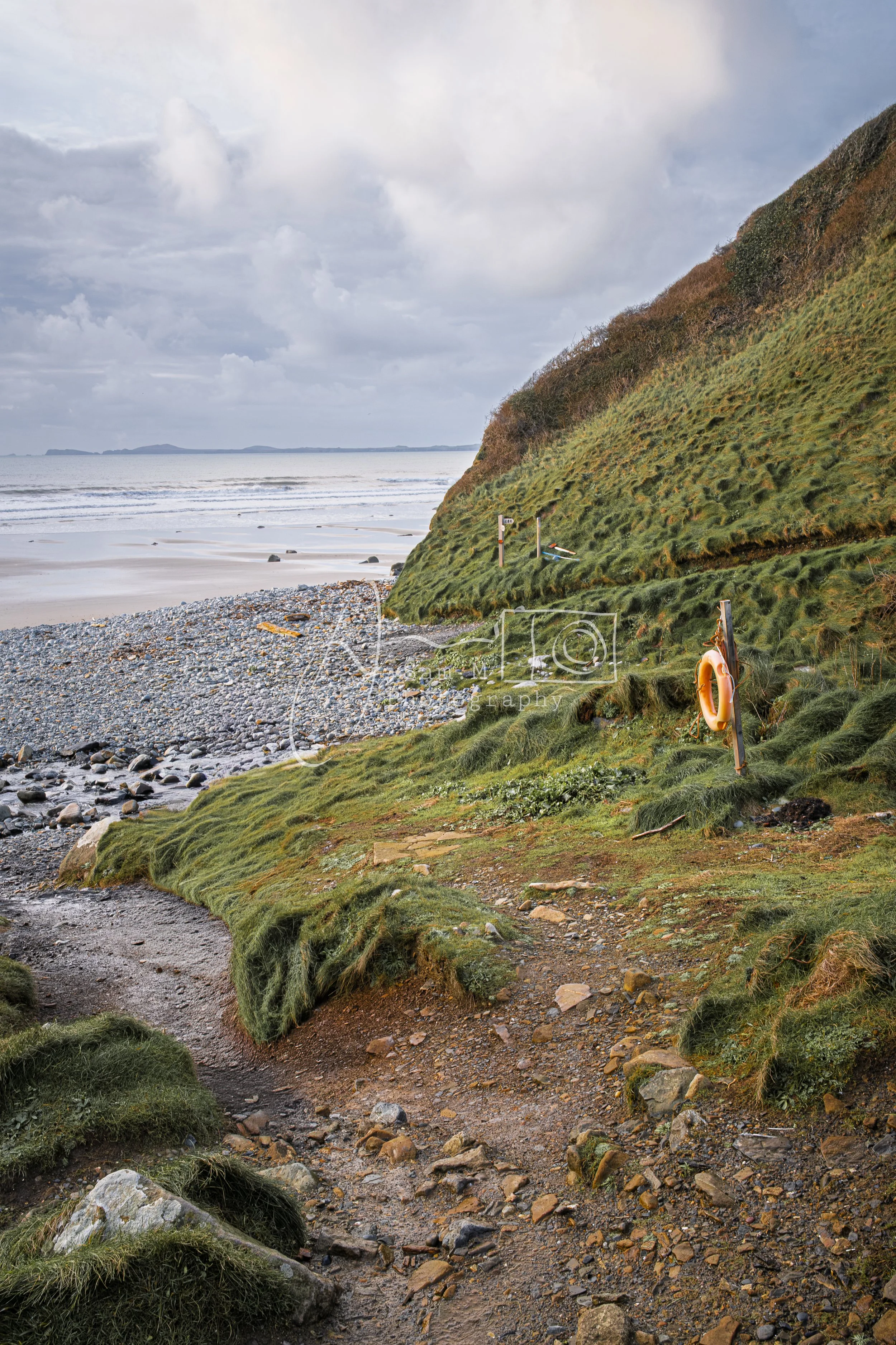 A rocky dirt trail along a grassy hillside leading to the beach, with life preservers attached to posts, overlooking the ocean on a partly cloudy day.
