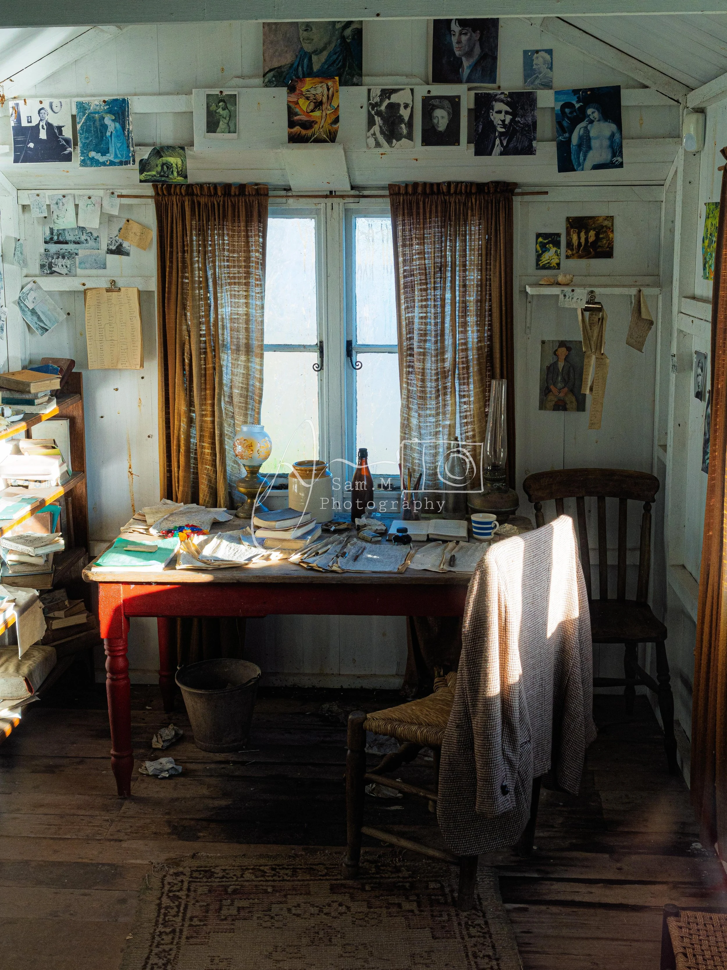 A cluttered wooden desk near a window with brown curtains, covered with papers, books, and a bottle. Two chairs, one with a coat or jacket draped over it, sit in front of the desk. The room has white wooden walls decorated with various photographs, p