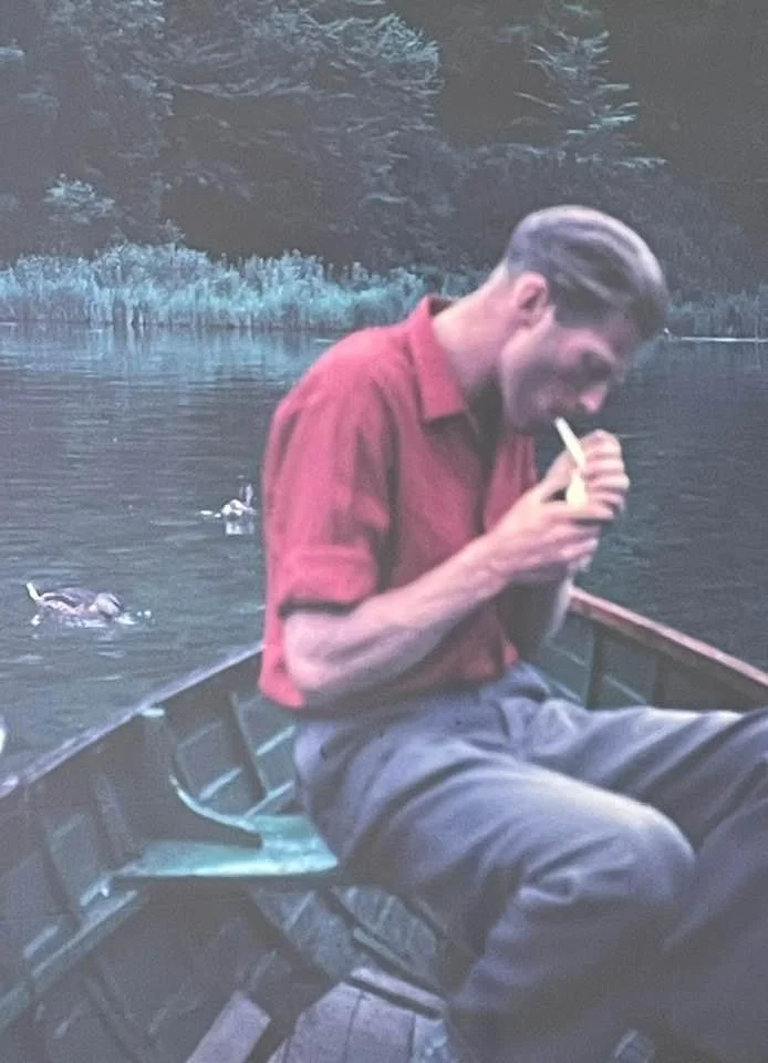 A man sitting in a boat on a lake, eating fries and playing on his phone, with ducks swimming nearby in the water.