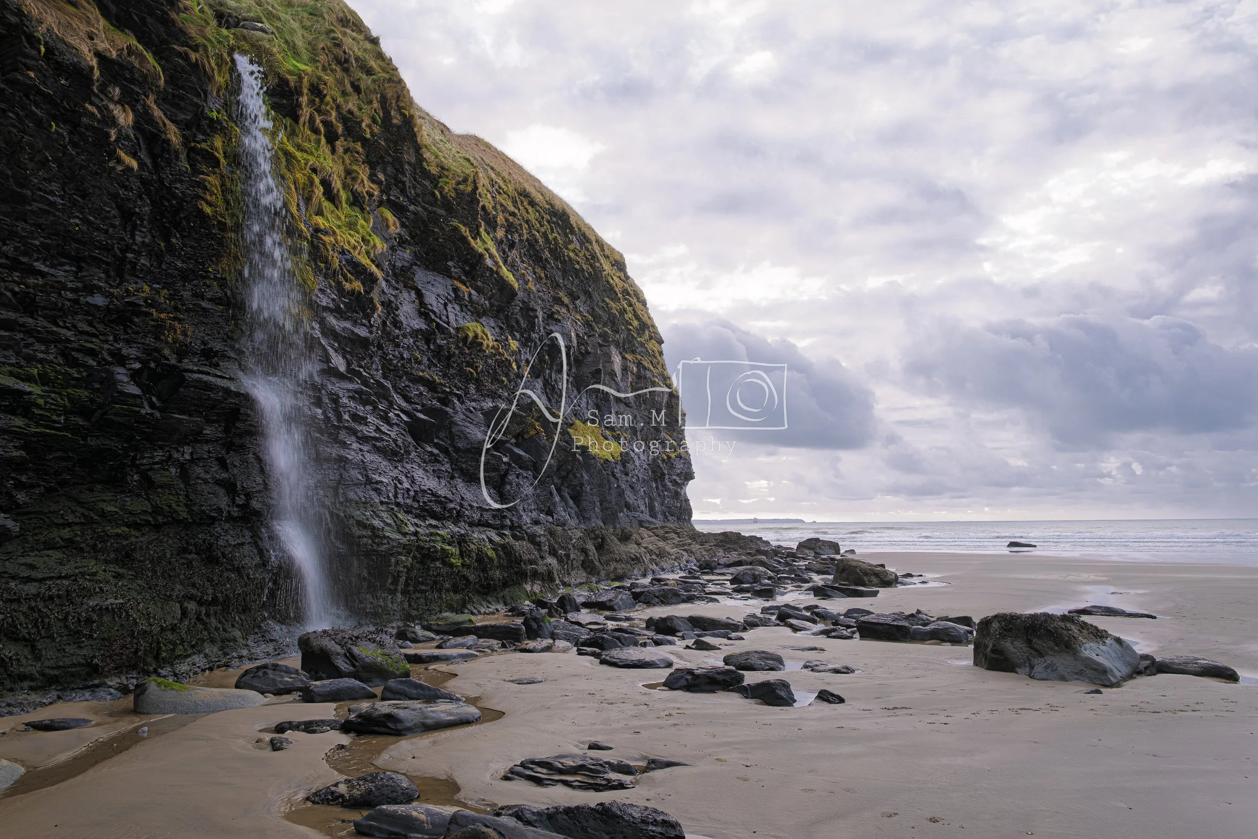 A beach with a rocky shoreline and a cliff on the left side, featuring a small waterfall flowing down the dark, moss-covered rocks. The sky is cloudy with a mix of sunlight and shadows, and the ocean waves are visible in the distance.