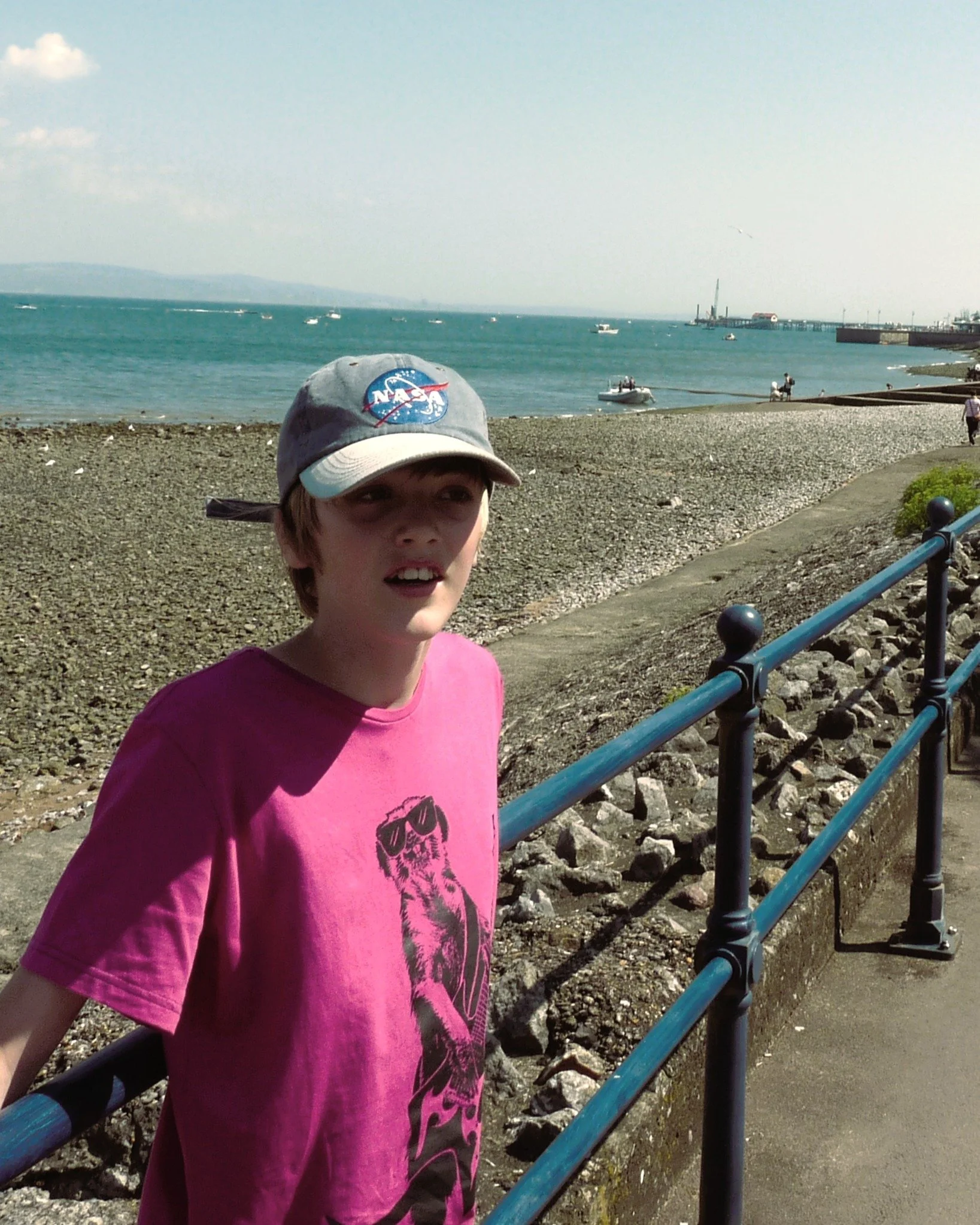 A young boy wearing a pink shirt and a gray NASA cap standing next to a blue metal railing on a seaside promenade, with the ocean, boats, and a pier visible in the background.