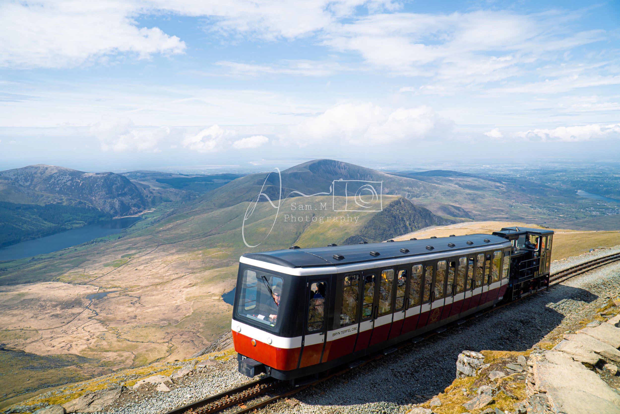 A cable car traveling along a mountain railway with a scenic view of rolling hills, a lake, and blue sky with clouds in the background.
