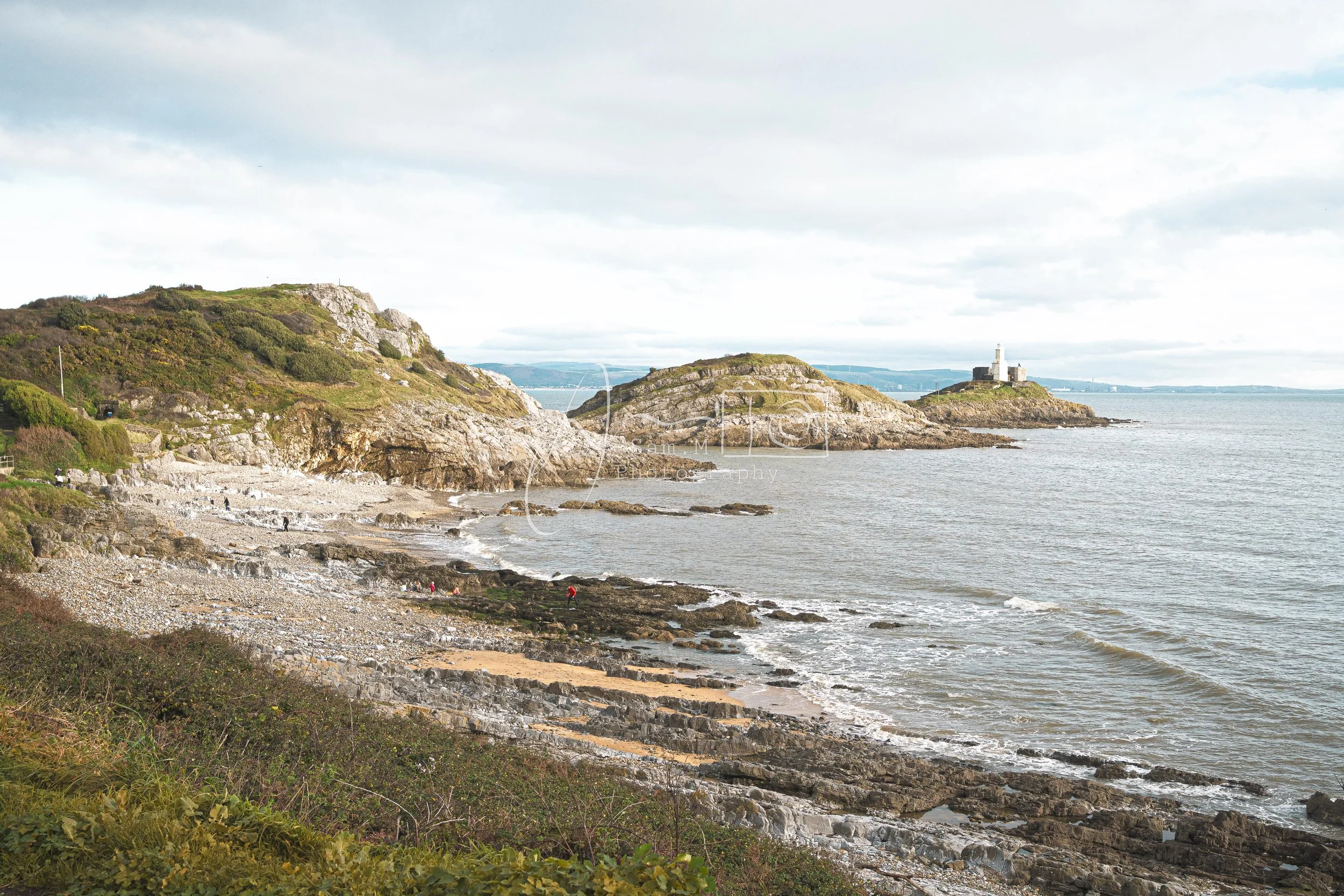 Coastal scene with rocky shoreline, green hills, lighthouse on small island, and calm waters under cloudy sky.
