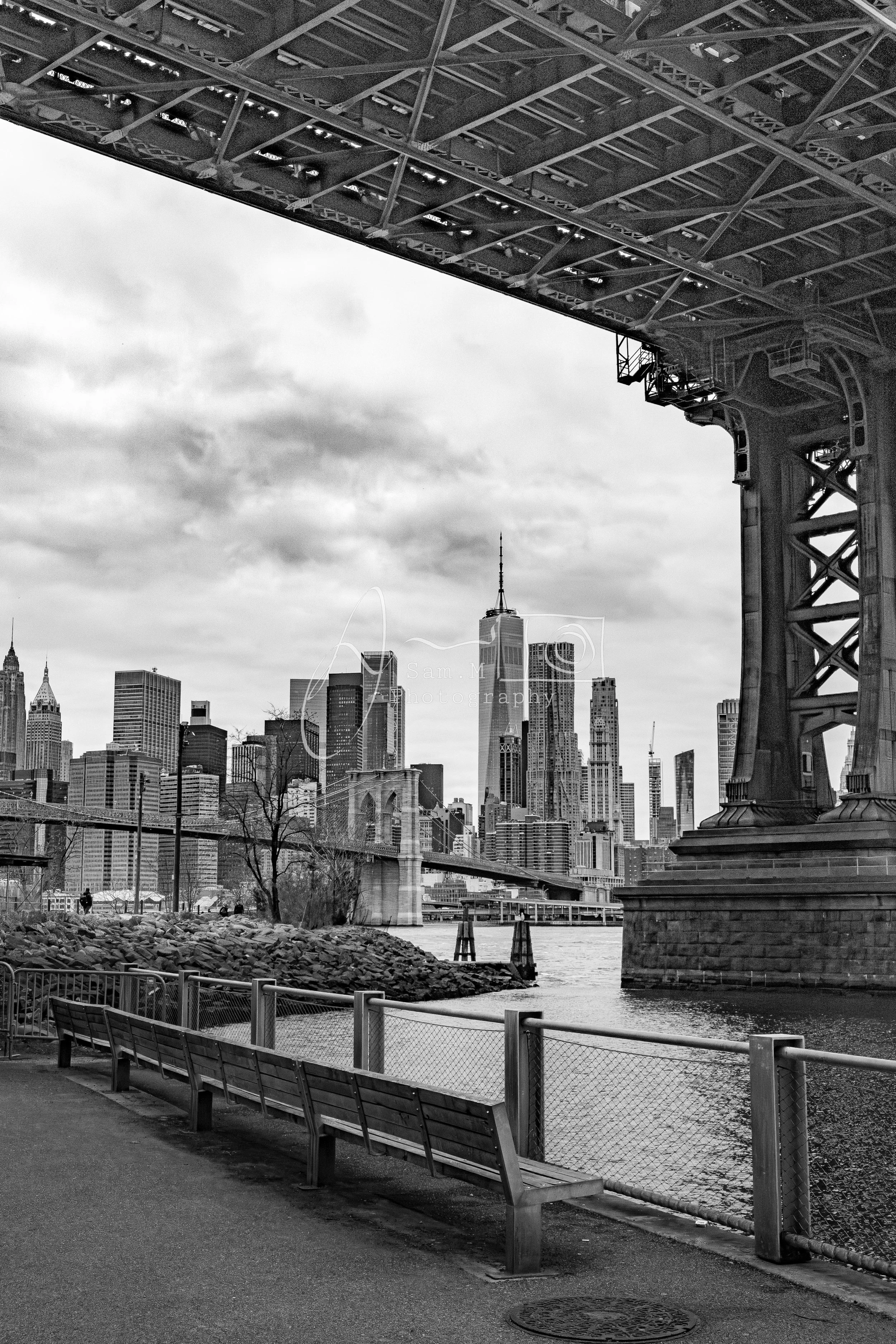 Black and white photo of the New York City skyline with the Brooklyn Bridge in the foreground, view from the Brooklyn Riverside Park with benches along the riverside.