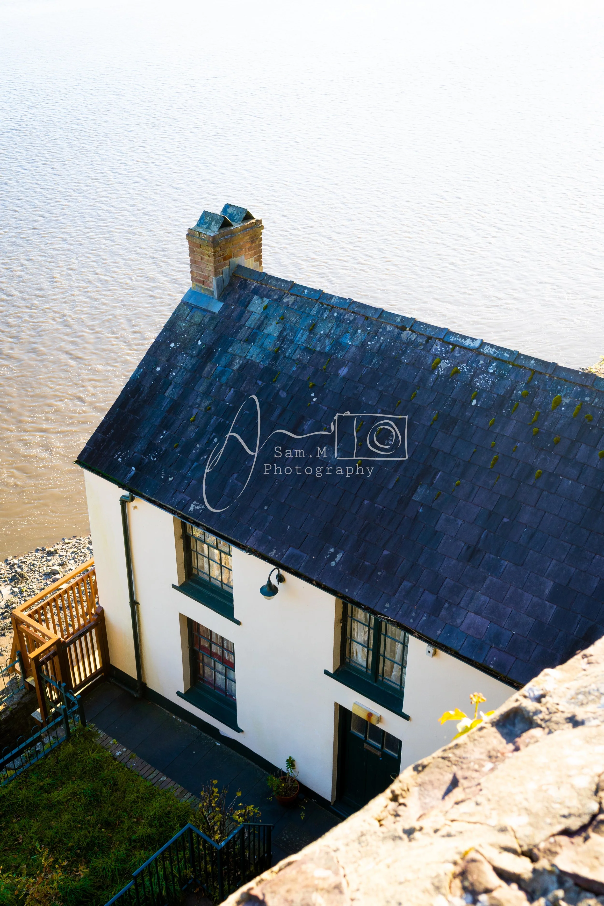 A white two-story house with a black door and window frames, a sloped slate roof, and a brick chimney, situated beside a body of water, viewed from an elevated angle.