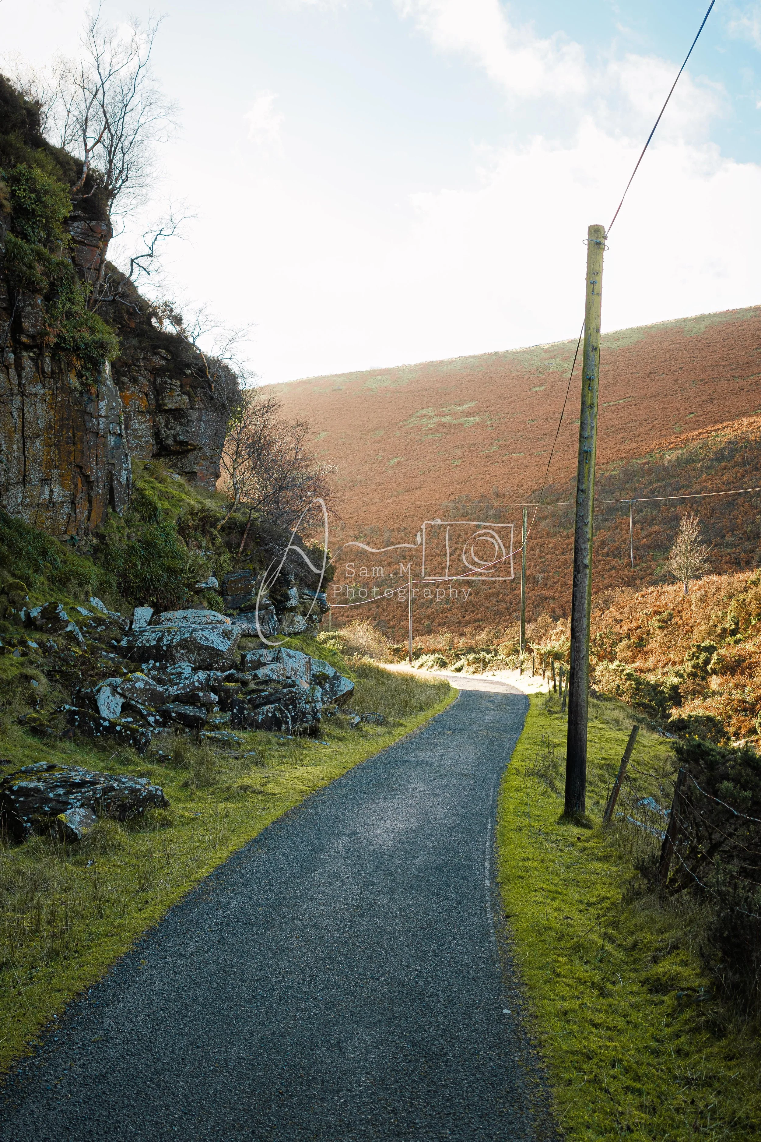 A narrow winding road through a rural landscape with green grass on both sides, a wooden utility pole with wires, rocky hillside on the left, and a hill covered in brown vegetation on the right, under a partly cloudy sky.