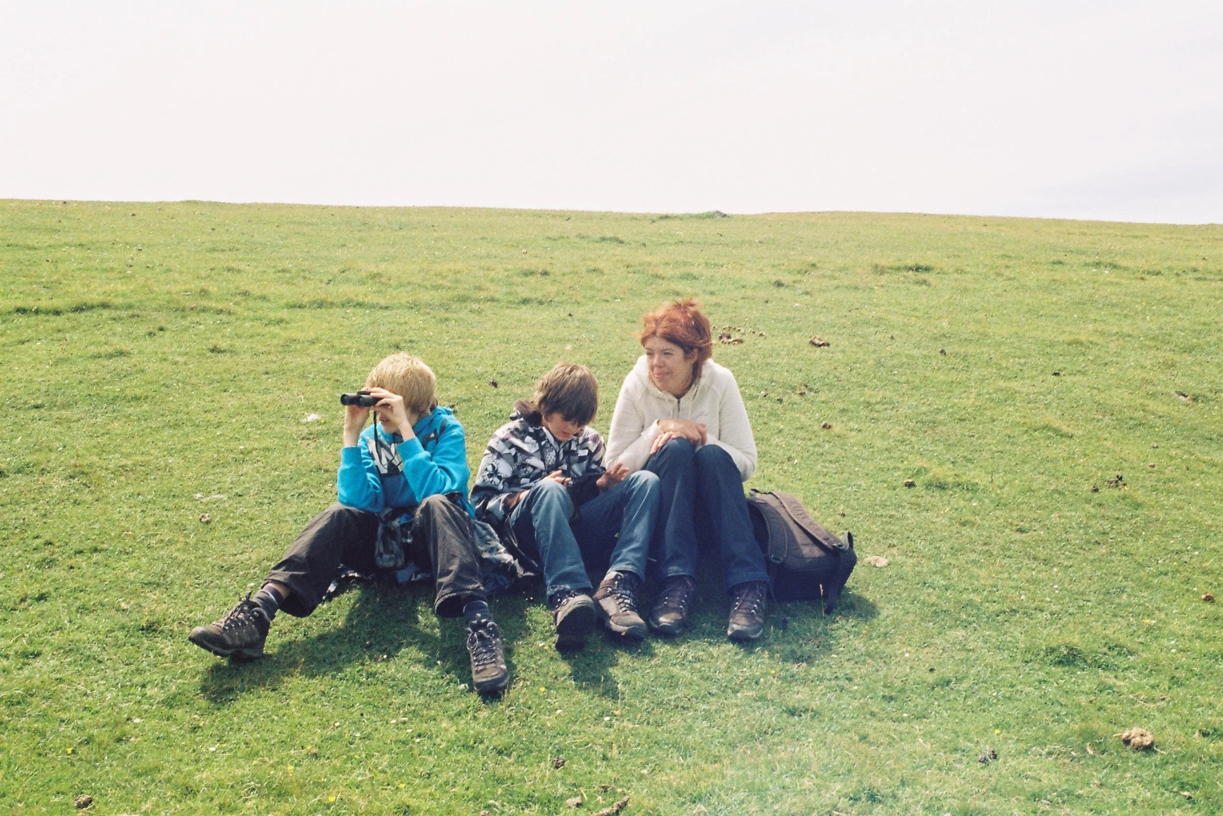 Three children sitting on green grass outdoors, with one child using binoculars, one looking at a device, and a woman sitting beside them, surrounded by backpacks.