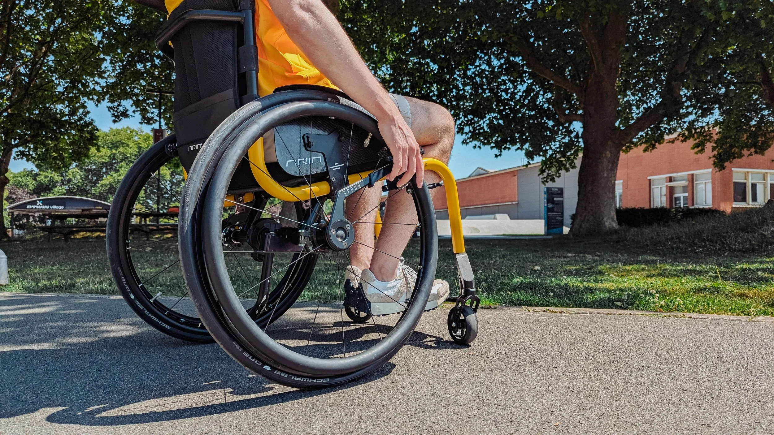 Close-up of a person in a wheelchair outdoors on a sunny day, touching the wheel, with trees and a brick building in the background.