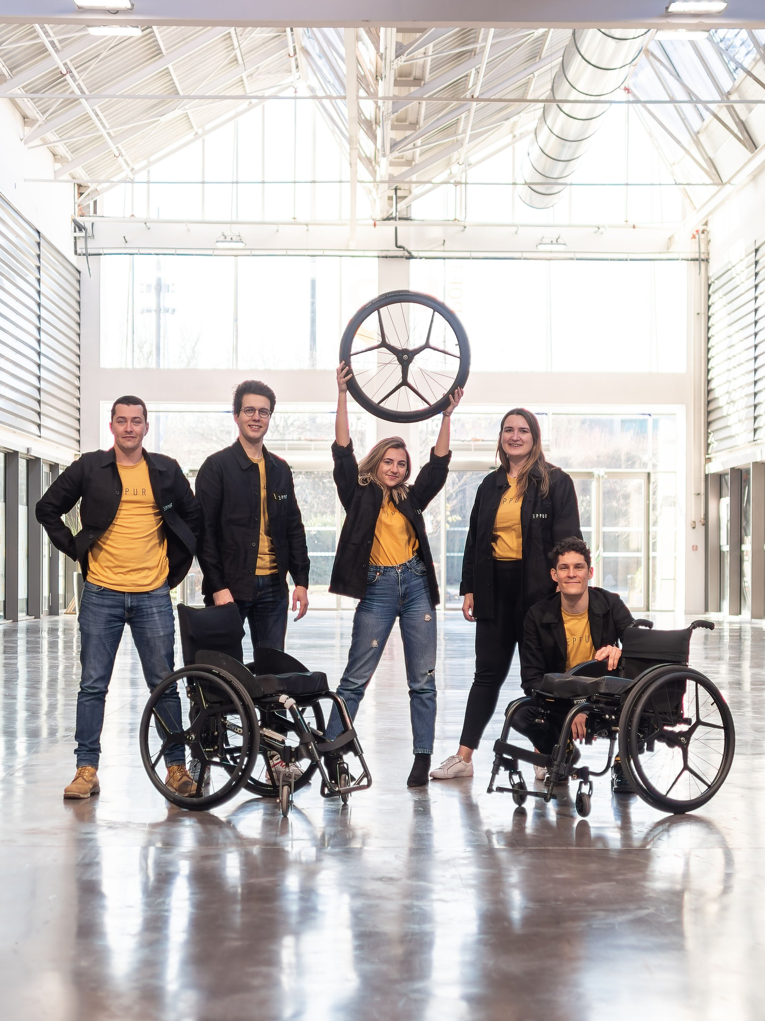 A group of five diverse young adults, including two in wheelchairs, posing inside a spacious, well-lit industrial-style building with large windows. One woman is holding an Eppur Dreeft wheelchair wheel with integrated braking above her head.
