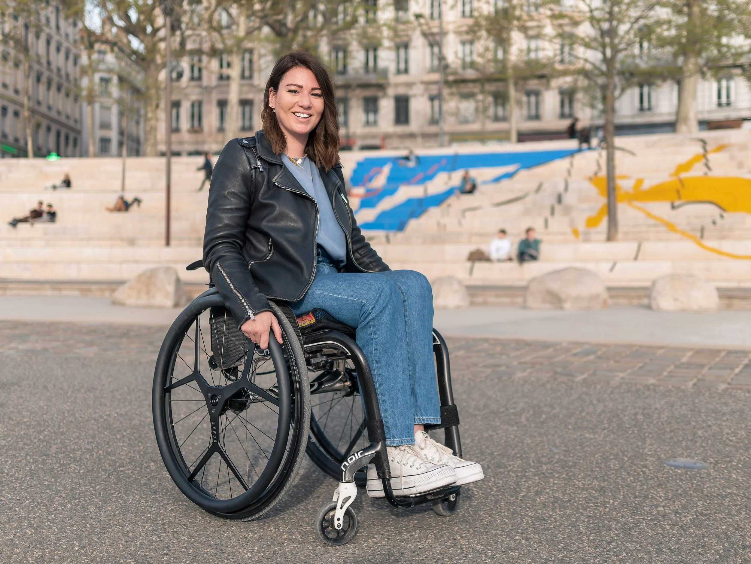 A young woman sitting in a wheelchair outdoors in an urban park, smiling and wearing a black leather jacket, blue jeans, and white sneakers. The background features a wide stone staircase with graffiti art and trees.